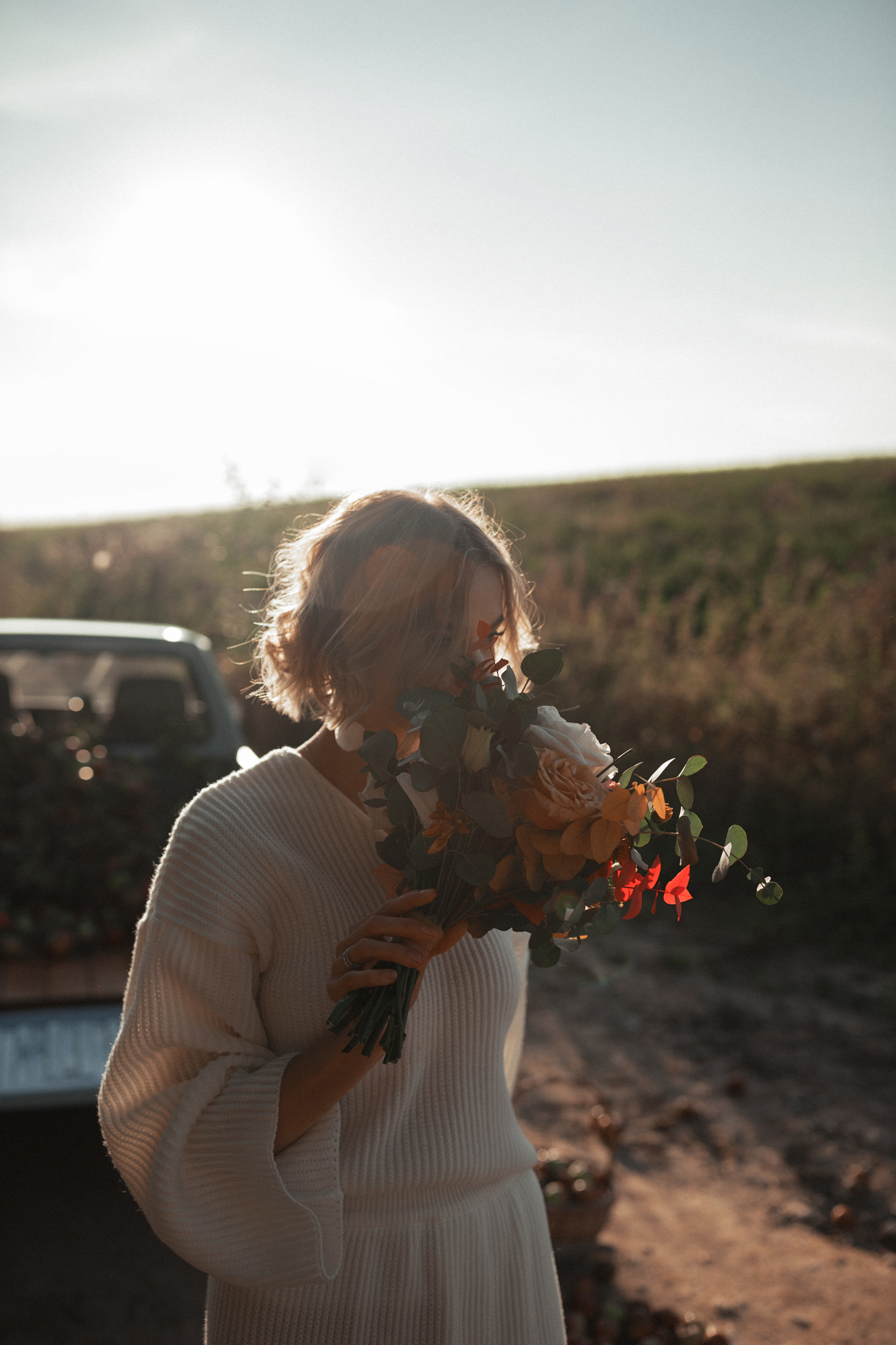 Невеста, стоя у машины нюхает букет красных цветов.