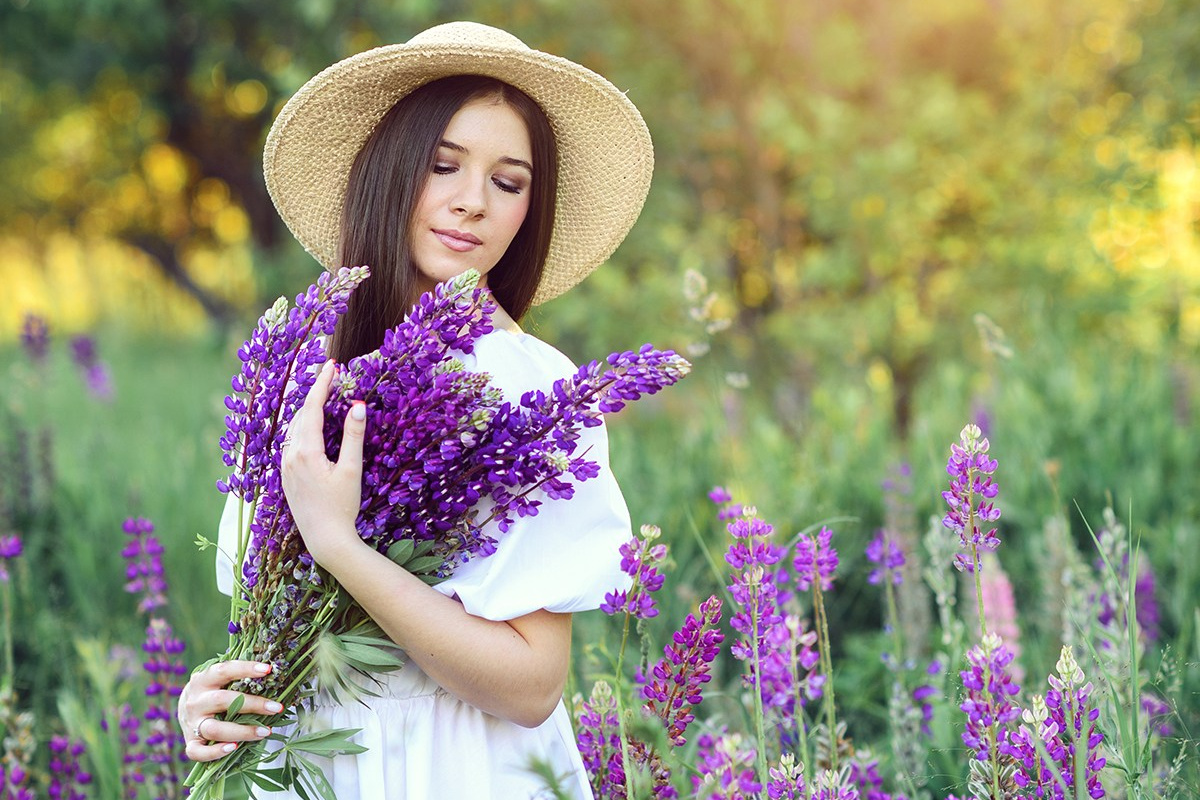 Красивая девушка на закате стоит в белом, легком платье и соломенной шляпке и держит в руках букет люпинов