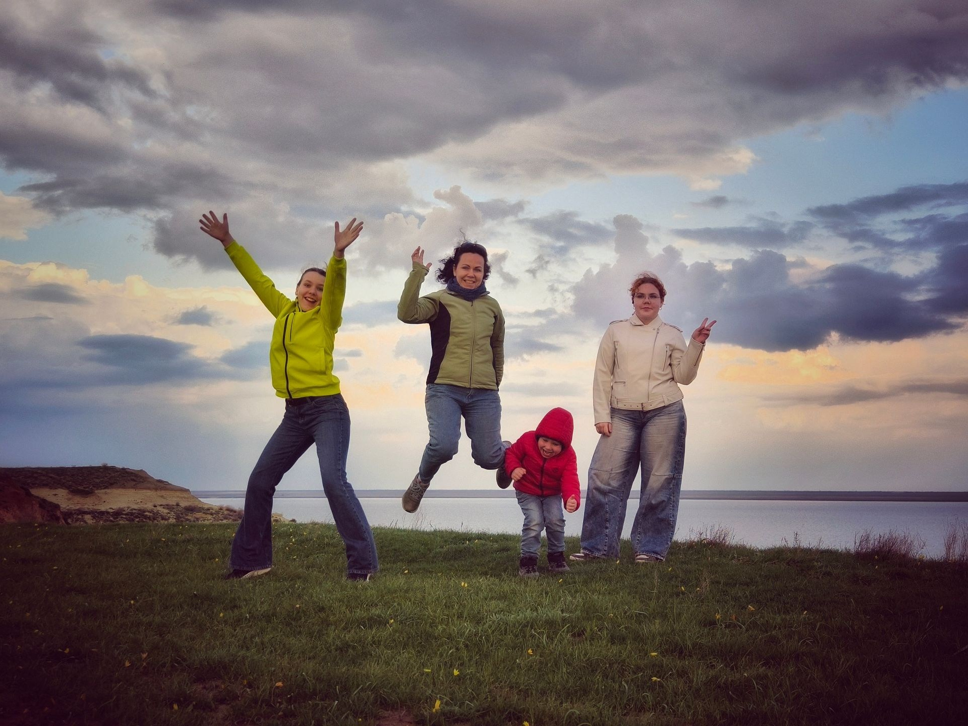 Семья радостно прыгает на траве у воды на фоне облачного неба