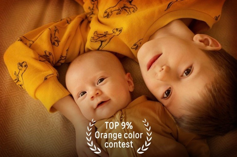 Мальчик обнимает свою сестру-младенца лежа на кровати в оранжевой одежде.