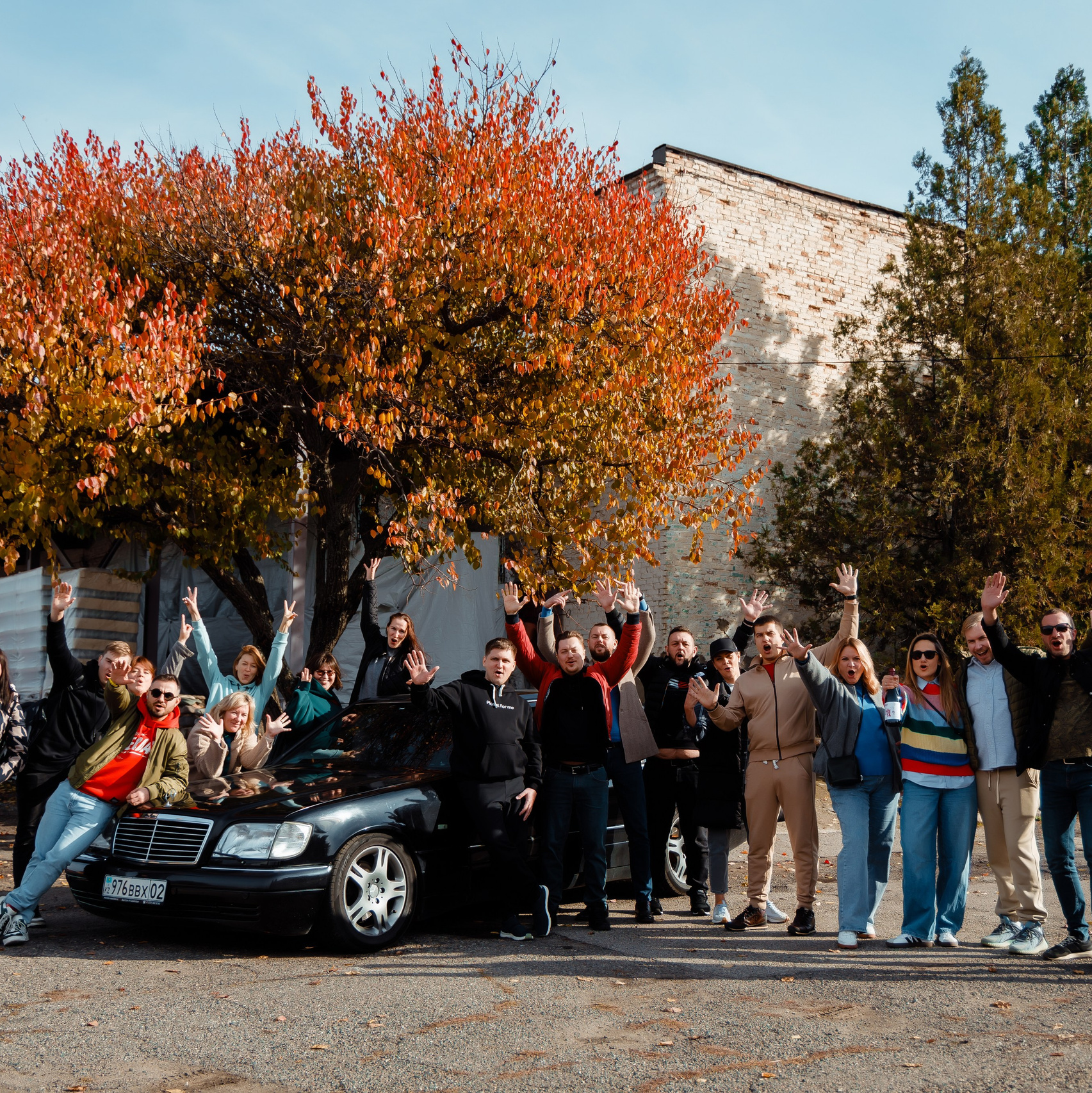 Группа сотрудников, стоящих на улице возле черного автомобиля на фоне ярко окрашенных осенних деревьев. Участники улыбаются и машут руками, создавая дружелюбную и позитивную атмосферу в корпоративном мероприятии. На заднем плане видны здания и зелёные деревья. Яркие цвета осенних листьев подчёркивают радостное настроение сцены.