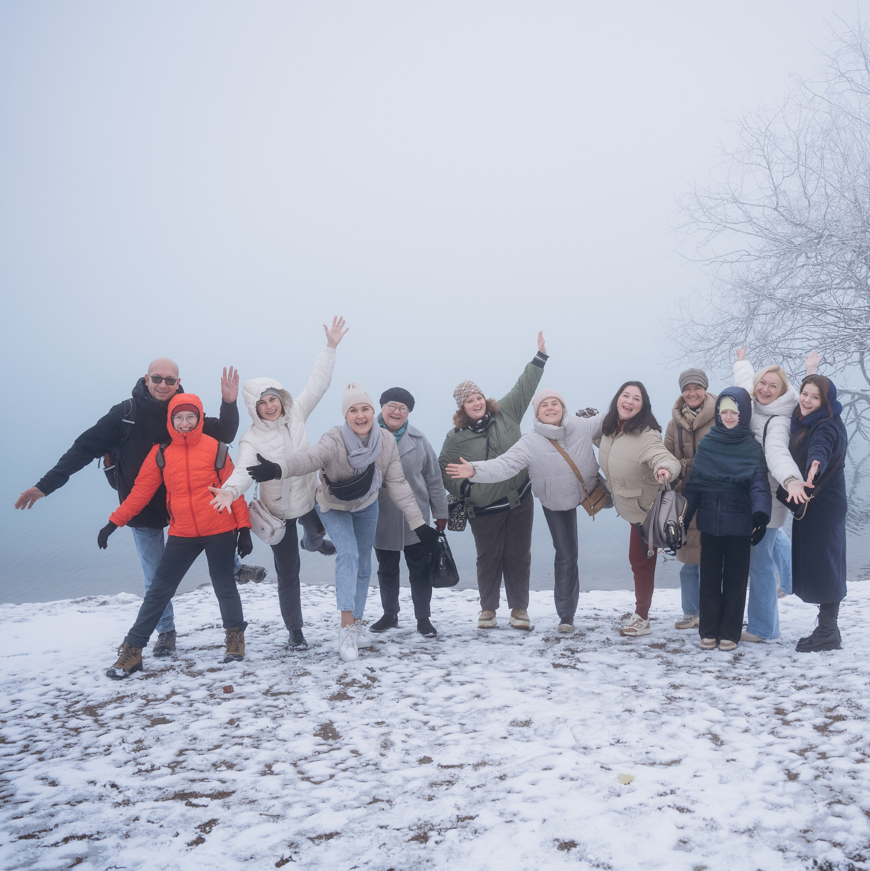 Группа людей, участников корпоративного тура Melon Travel на озеро Иссык, одетых в зимнюю одежду, позирует на фоне заснеженного пейзажа. На земле лежит снег, деревья покрыты инеем, а на заднем плане виднеется туман, создающий атмосферу спокойствия и загадочности. Участники группы улыбаются и выглядят счастливыми.