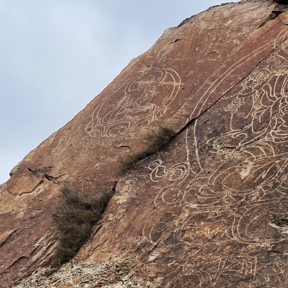 Высокий скалистый утес в пустынном ландшафте возвышается под ярким голубым небом с небольшими облаками. Поверхность скалы изрезана эрозией, придавая ей причудливую форму. Вокруг видна сухая земля с небольшими участками белого налета, возможно, из-за снега или соли. На заднем плане — более низкие скальные образования Чарынского каньона Казахстана.