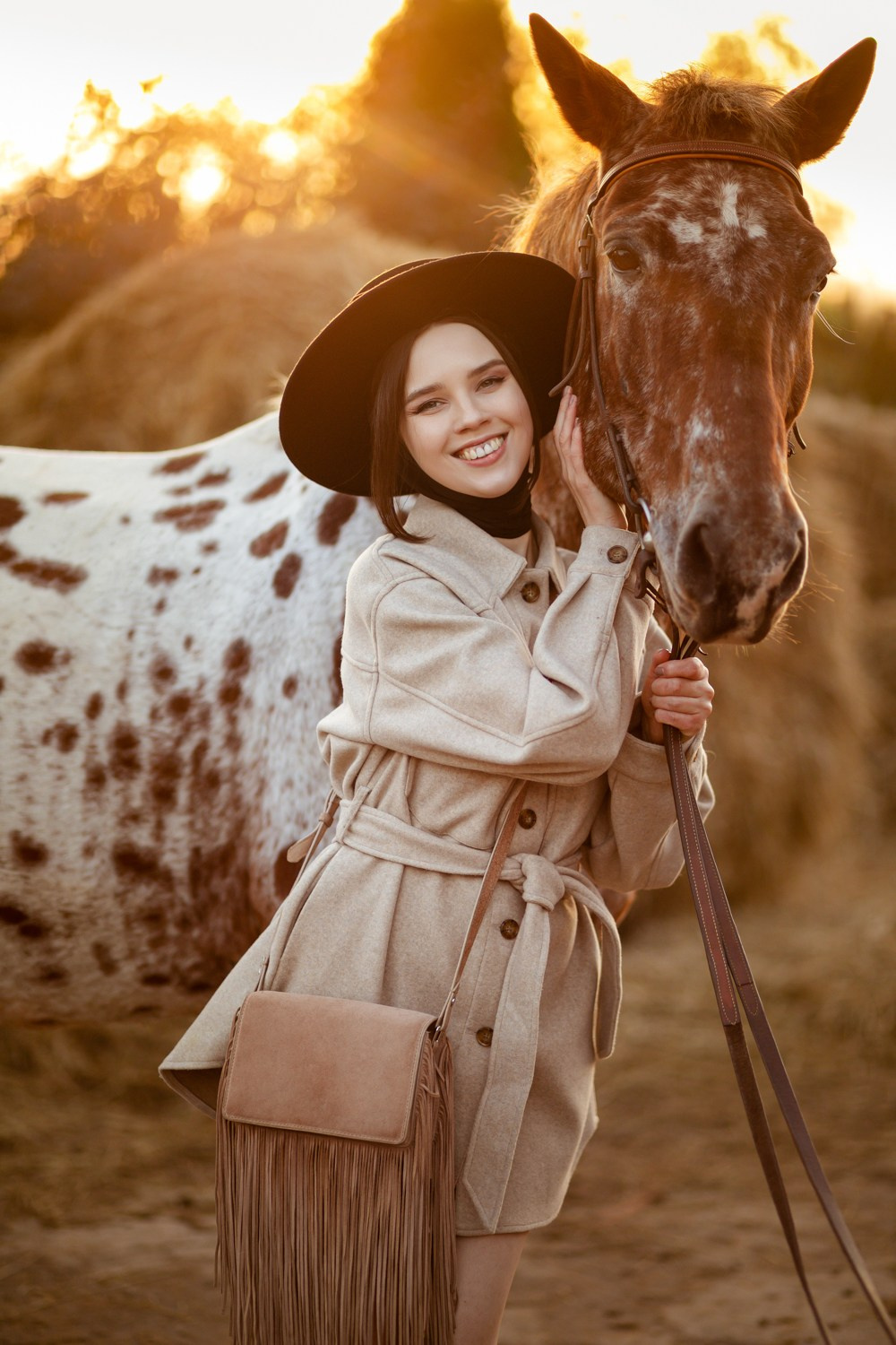 Девушка в бежевом пальто держит лошадь и улыбается
