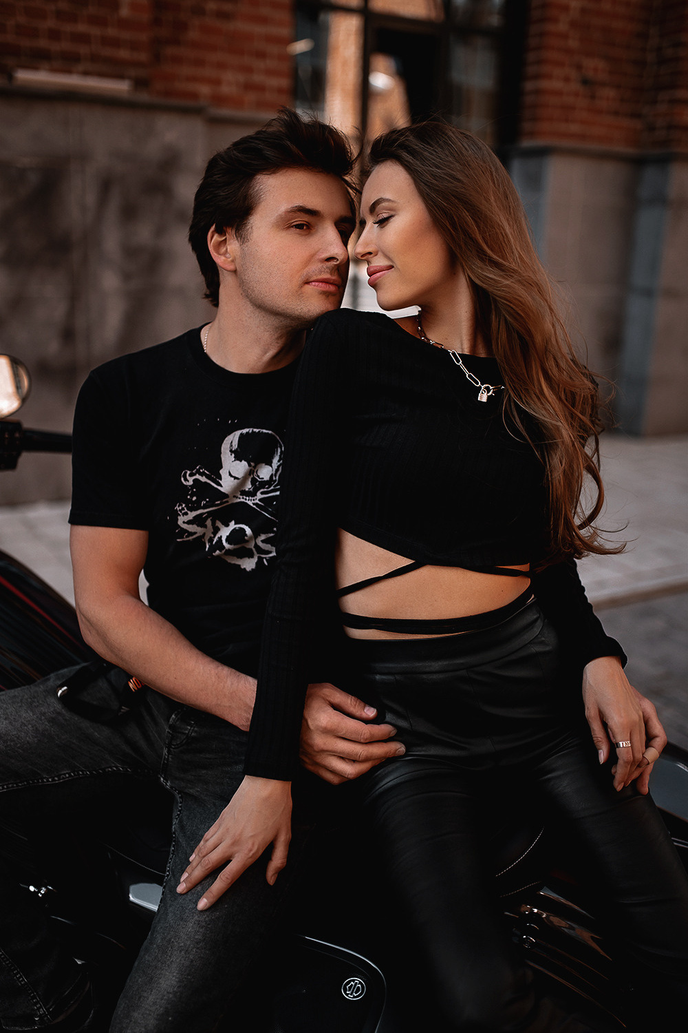 Пара, сидящая на мотоцикле. Мужчина одет в черную футболку с рисунком, а женщина в черный топ с длинными рукавами и кожаные брюки. Они смотрят друг на друга с улыбками, создавая романтическую атмосферу. 