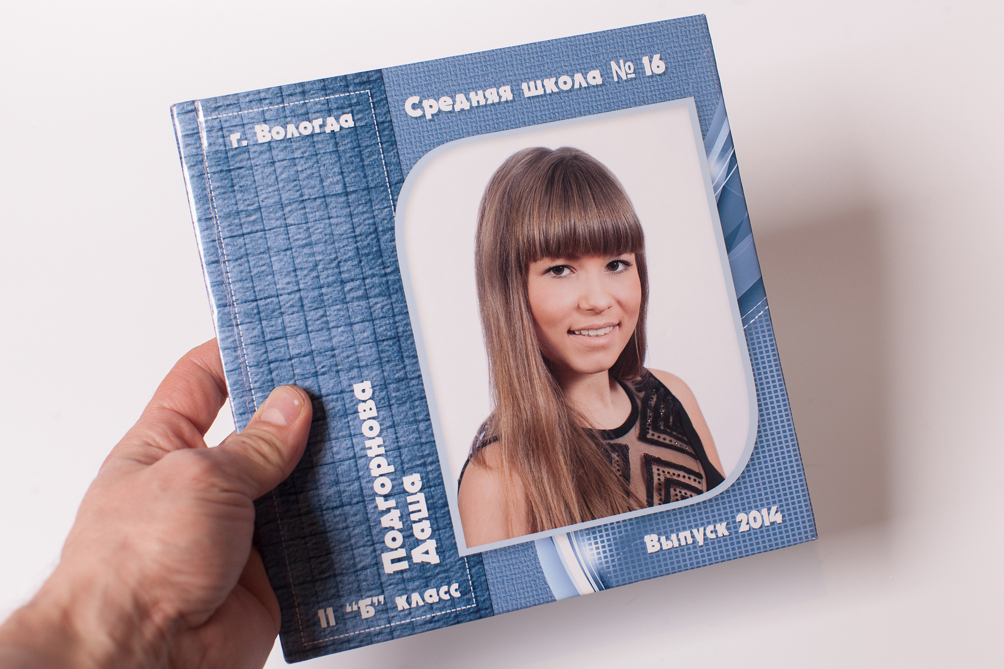 Рука держит синюю выпускную фотокнигу с портретом выпускницы 11 класса на обложке на светло-сером фоне.