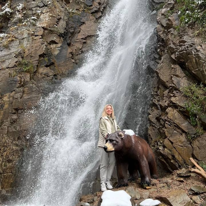 Пейзаж, напоминающий лунный, с извилистой рекой, окруженной густой зеленью и деревьями на фоне пустынных скалистых гор Лунного каньона. Солнечный день с ярко-голубым небом, украшенным небольшими облаками, подчеркивает природную красоту местности. Река Чарын образует небольшие островки, покрытые травой и кустарником.