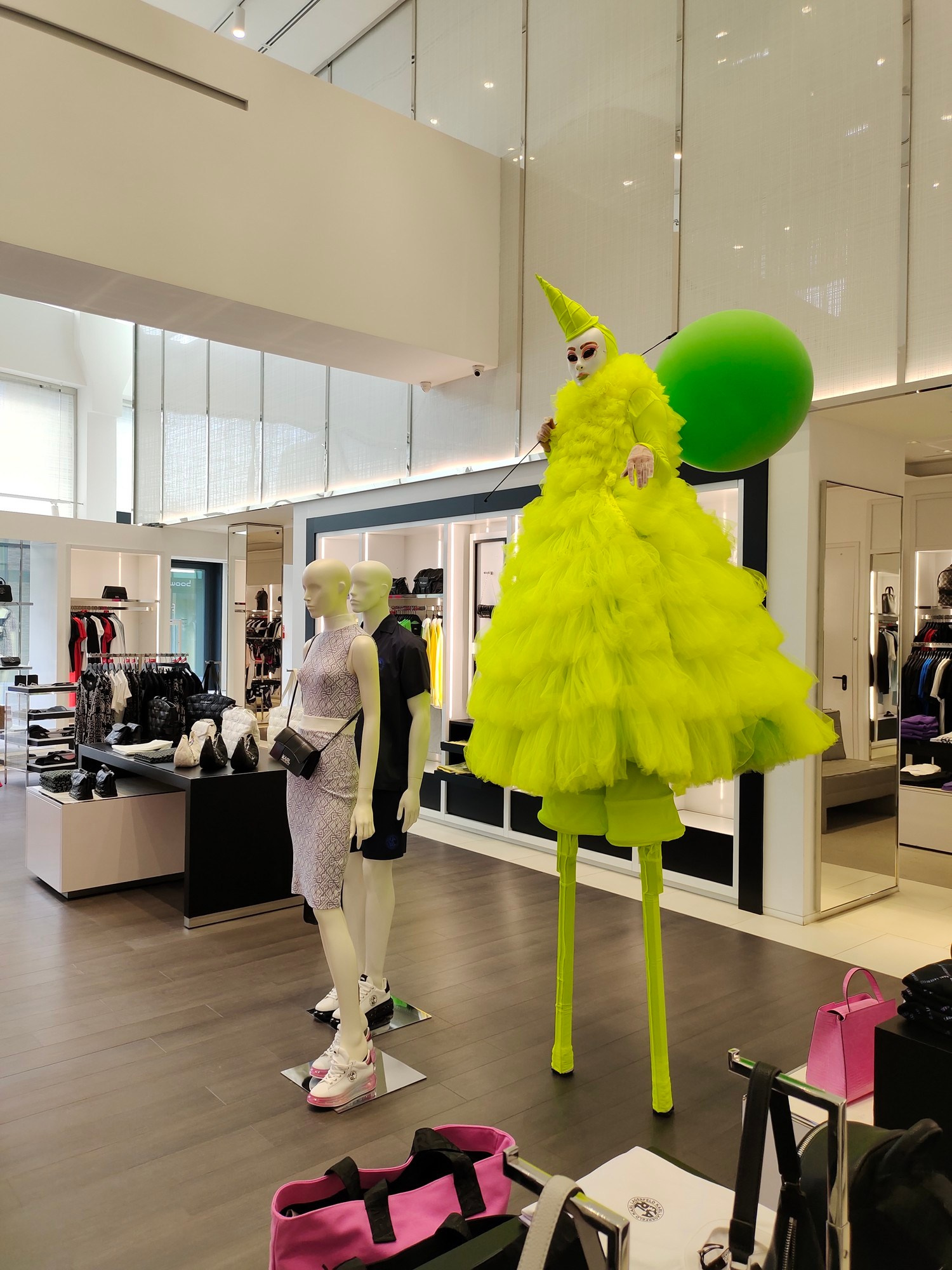 Ходулист ходит внутри магазина в зелёном платье и с зелёным шаром