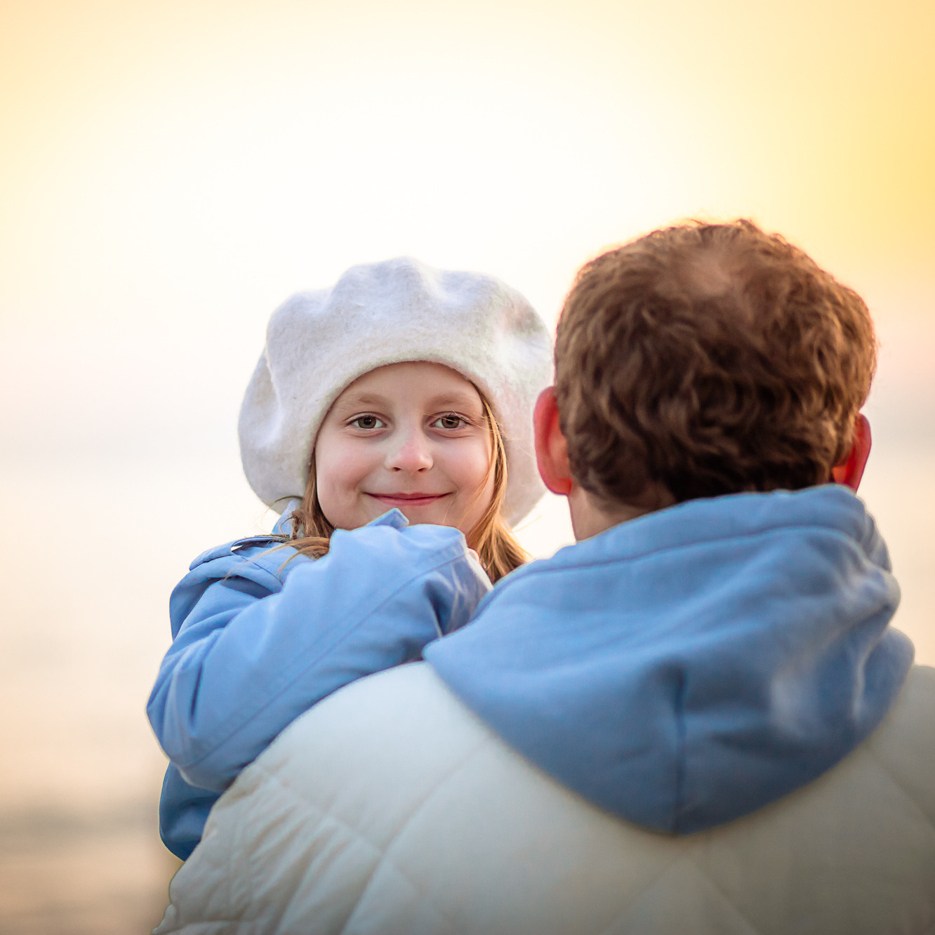Девочка у папы на руках смотрит через его плечо на закатном солнце