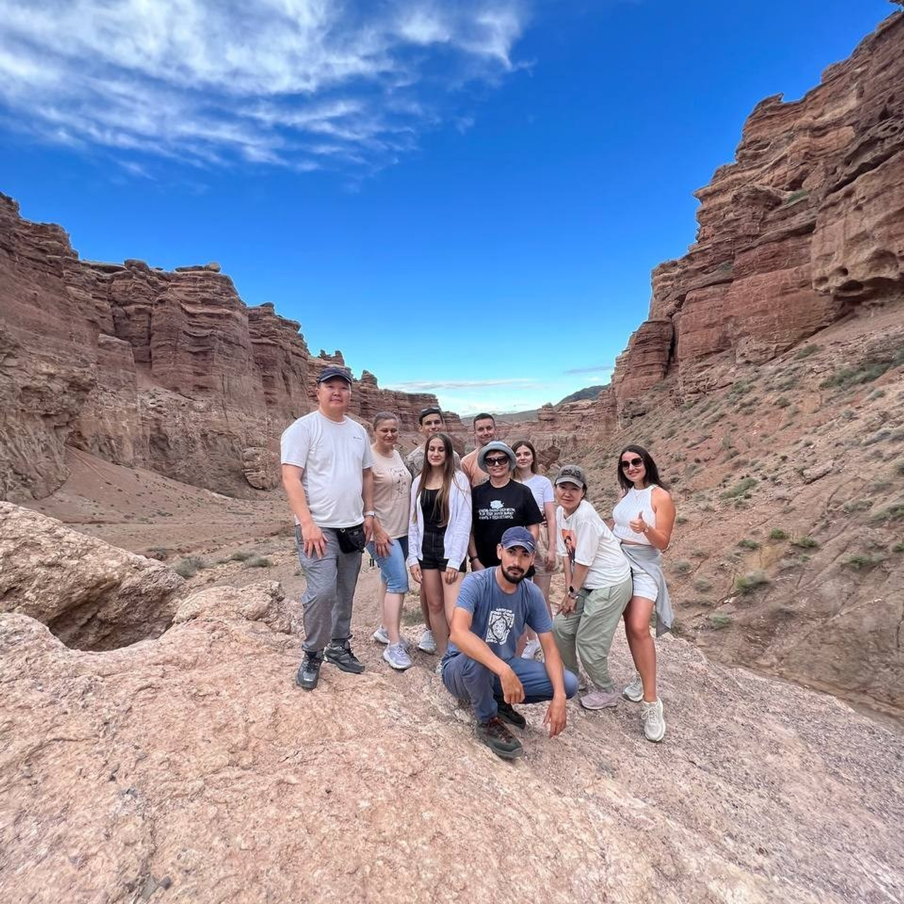 Группа людей позирует на фоне Долины Замков под ярким голубым небом. Они стоят и сидят на светло-коричневых камнях, одеты в удобную туристическую одежду. Атмосфера дружеская и лёгкая, а ландшафт вокруг подчёркивает величие Чарынского каньона.