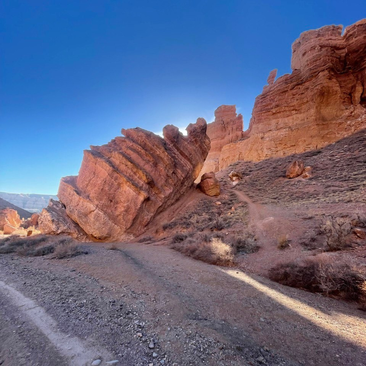 Участок Чарынского каньона с массивной наклонной скалой, освещённой солнцем, которое спряталось за ней. Оранжево-красные оттенки скал Долины Замков контрастируют с насыщенно-синим небом. Песчаная дорога и сухая растительность подчёркивают суровость и естественную красоту ландшафта.