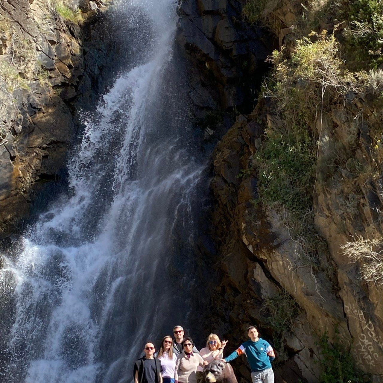 Группа из шести человек позирует у подножия Медвежьего водопада, который спускается с отвесных скал. В центре композиции также находится скульптура медведя. Каменистая местность, обрамлённая зеленью и солнечным светом, подчёркивает природную красоту места. Водопад выглядит мощным и освежающим на фоне синего неба.