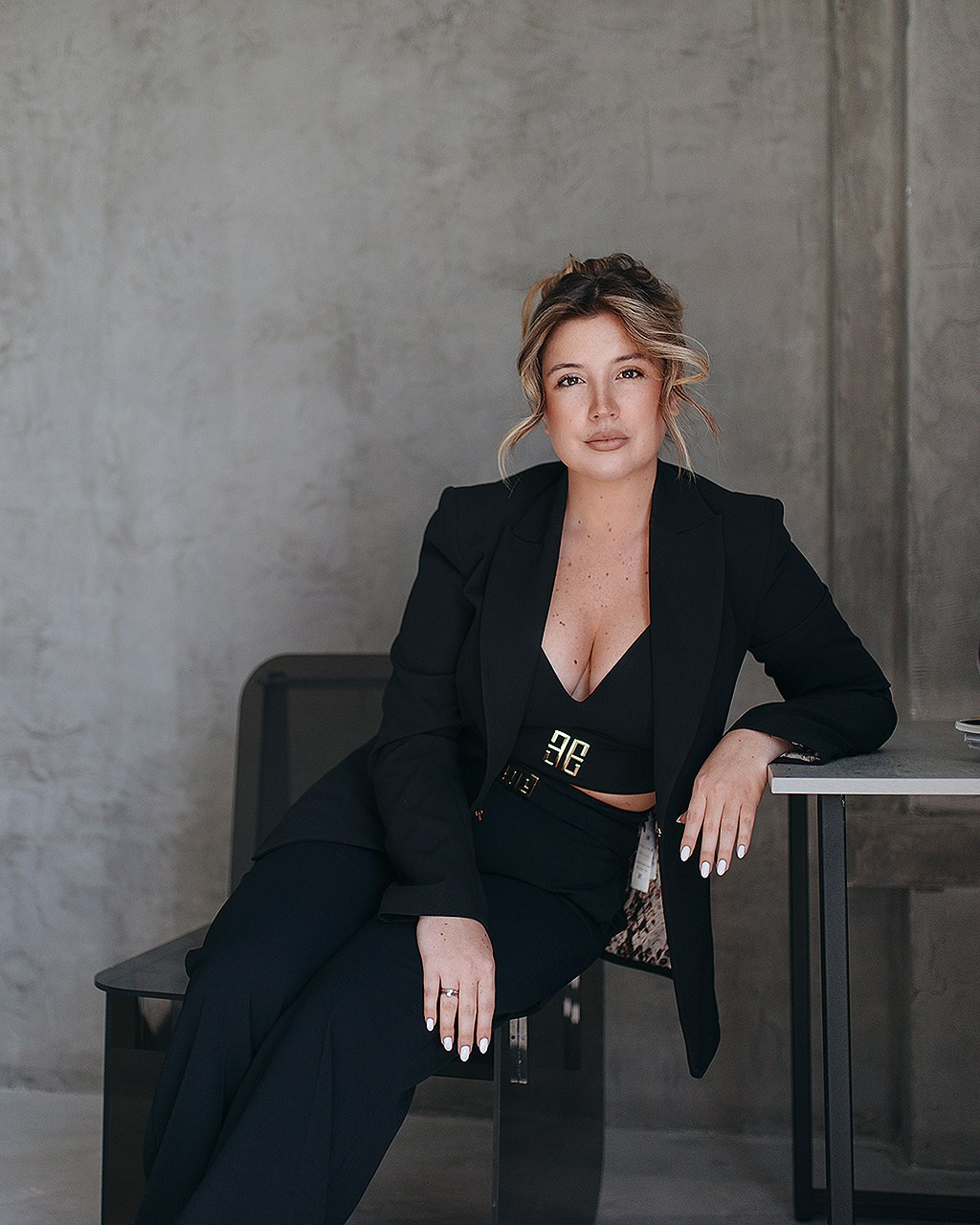 Красивая девушка с собранными волосами в деловом костюме сидит на стуле, опираясь локтем на стол. Студийная фотосессия на сером фоне.