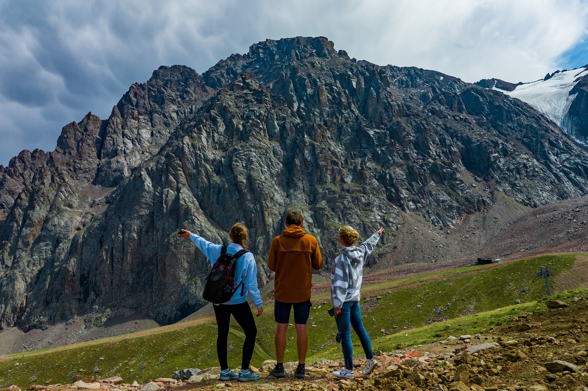 Человек позирует на фоне бирюзового озера Иссык в районе Алматы и гор. Он стоит на большом камне, одет в ярко-зеленую толстовку, черные штаны и светлую обувь, а также оранжевую панаму. В руках держит очки. За ним виднеются деревья, лесистые склоны и горные вершины под облачным небом. Атмосфера спокойная и природная.