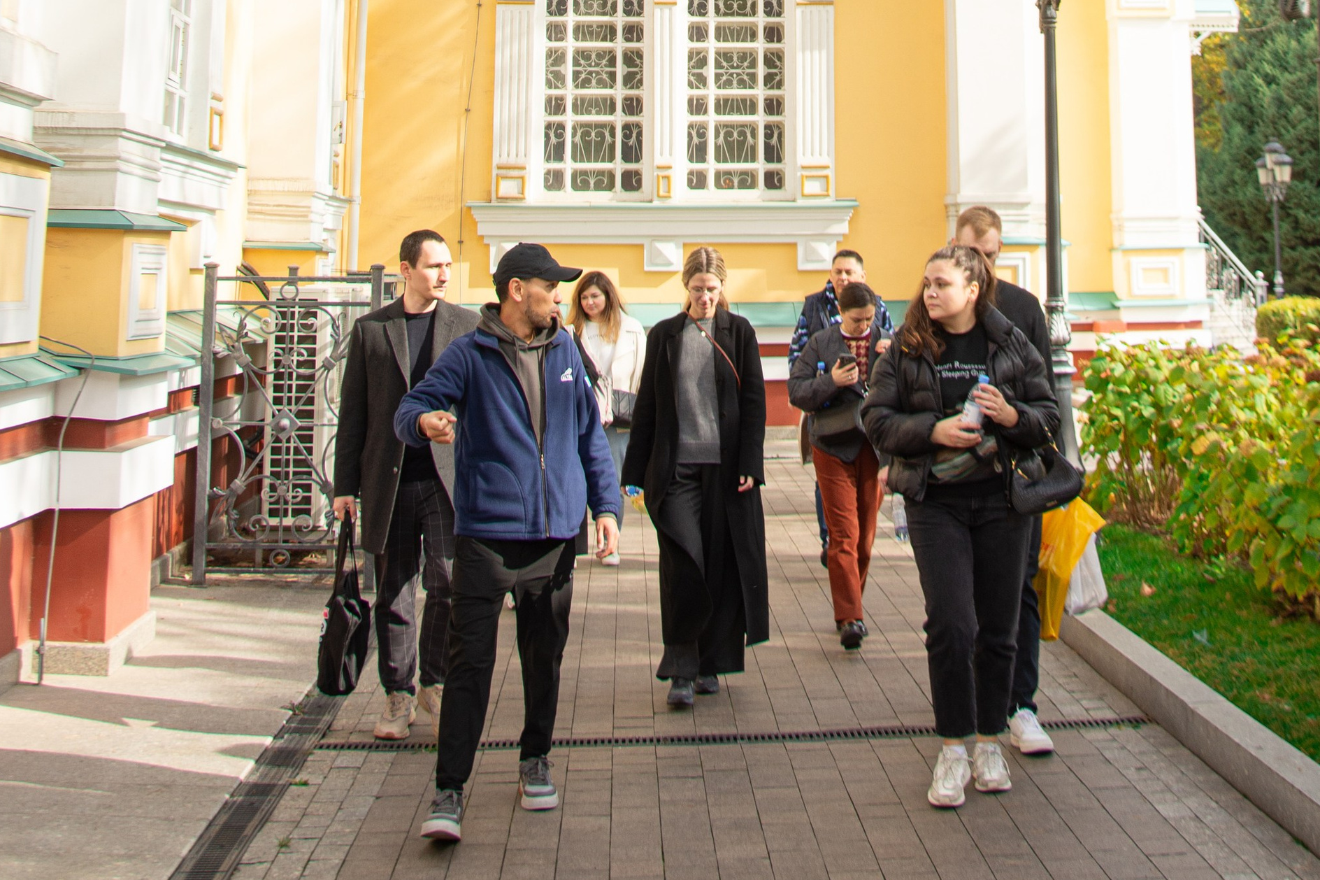 Группа из четырёх человек улыбается и позирует на фоне деревянной арки с надписью "Медвежий водопад". Люди выглядят счастливыми, одеты в светлую и спортивную одежду, очевидно, находятся в походе. За аркой видны склоны с зеленью и каменистая тропа. Атмосфера весёлая и дружелюбная, подчеркивает любовь к природе и приключениям.