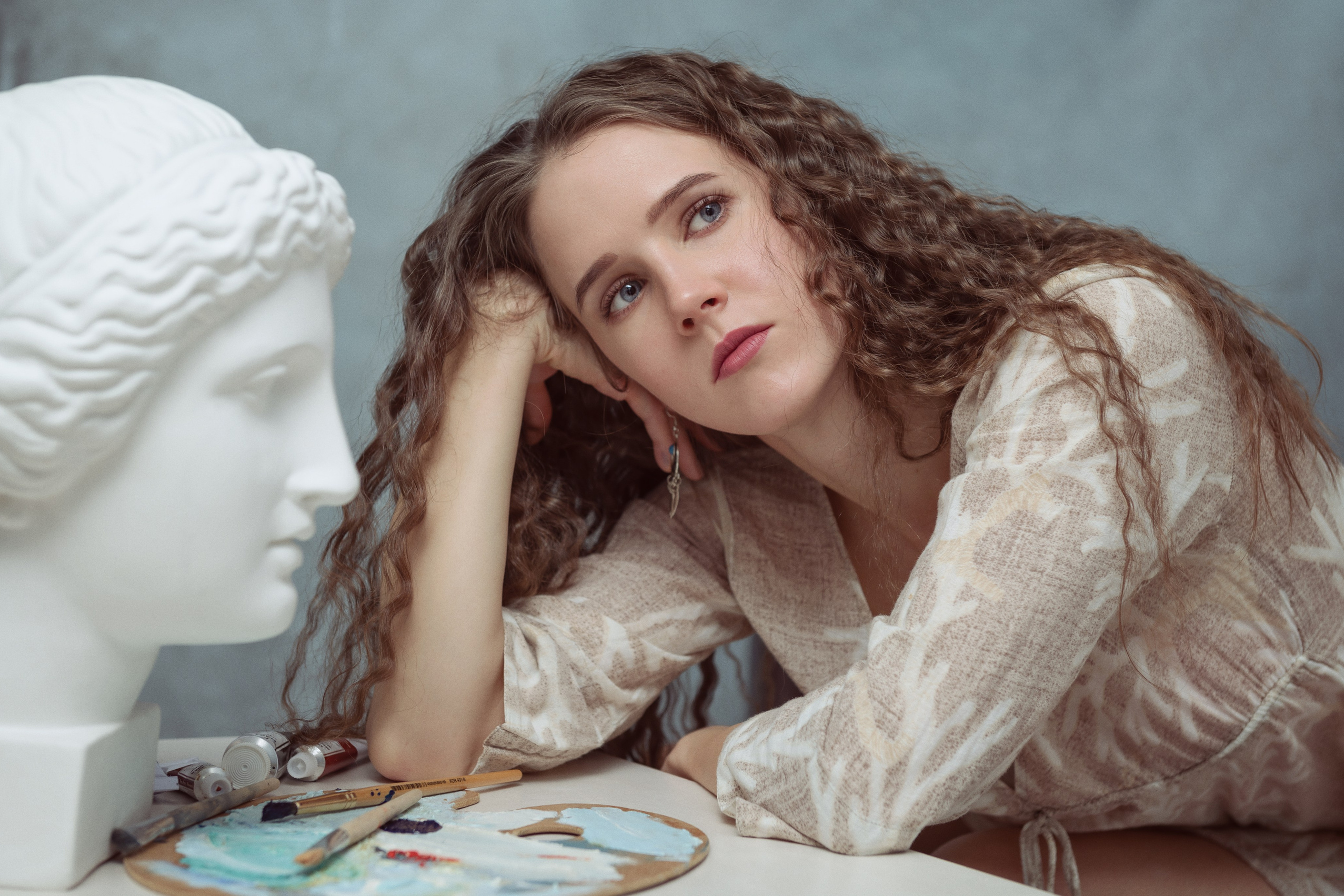 Художественное фото девушки с кудрявыми волосами, она предстает в образе художницы, которая задумчиво сидит рядом с гипсовой головой Музы