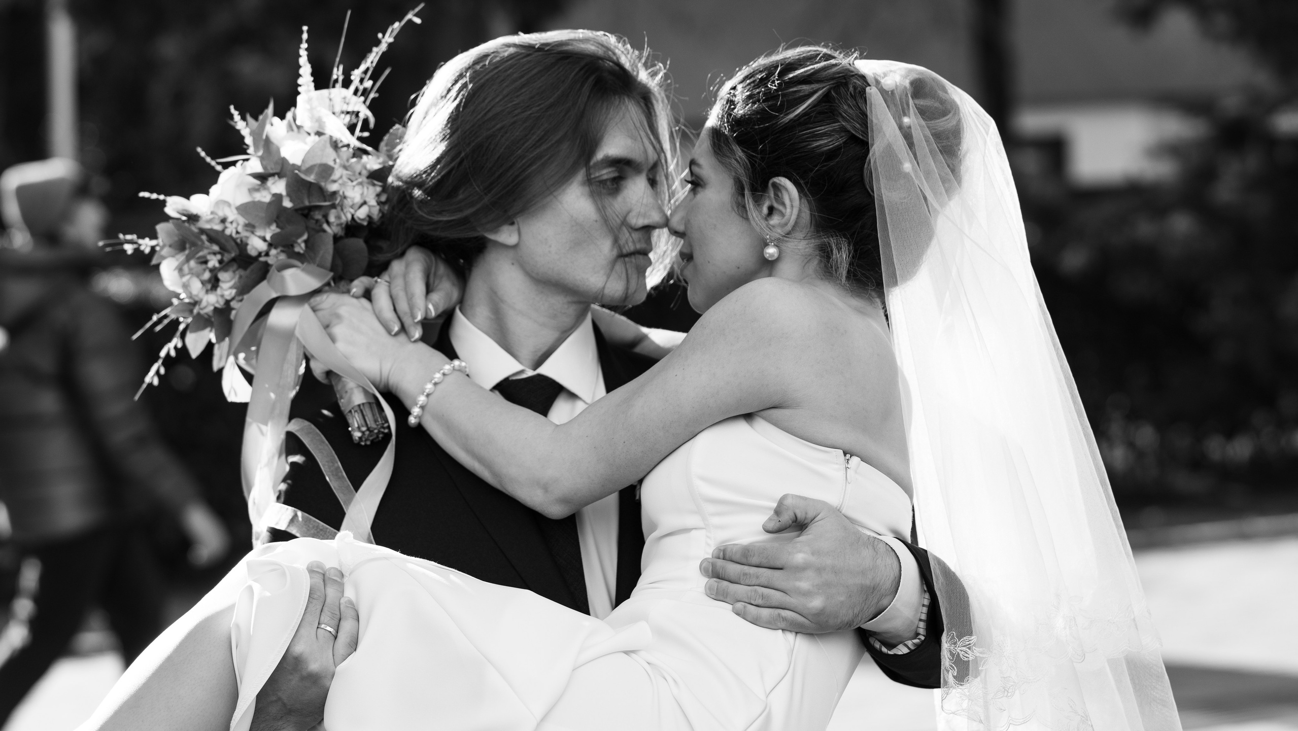 Это черно-белая фотография жениха и невесты в день их свадьбы. Жених целует невесту, неся ее на руках. Невеста одета в белое свадебное платье и фату. Жених одет в черный смокинг и бутоньерку из белых роз. Невеста держит букет белых цветов. Фон размыт.