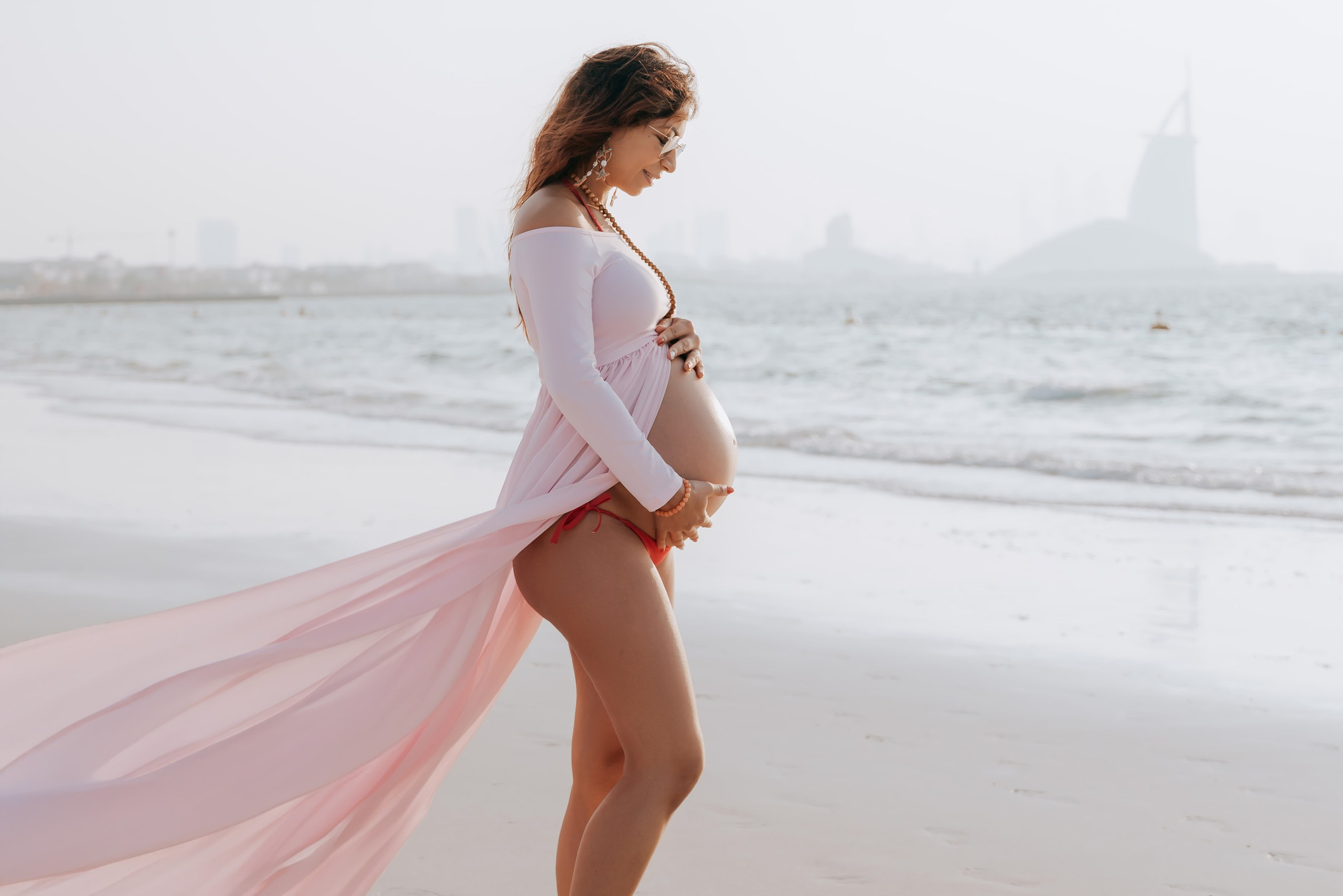 A pregnant woman in a flowing pink dress stands on a beach, capturing the serene beauty of maternity