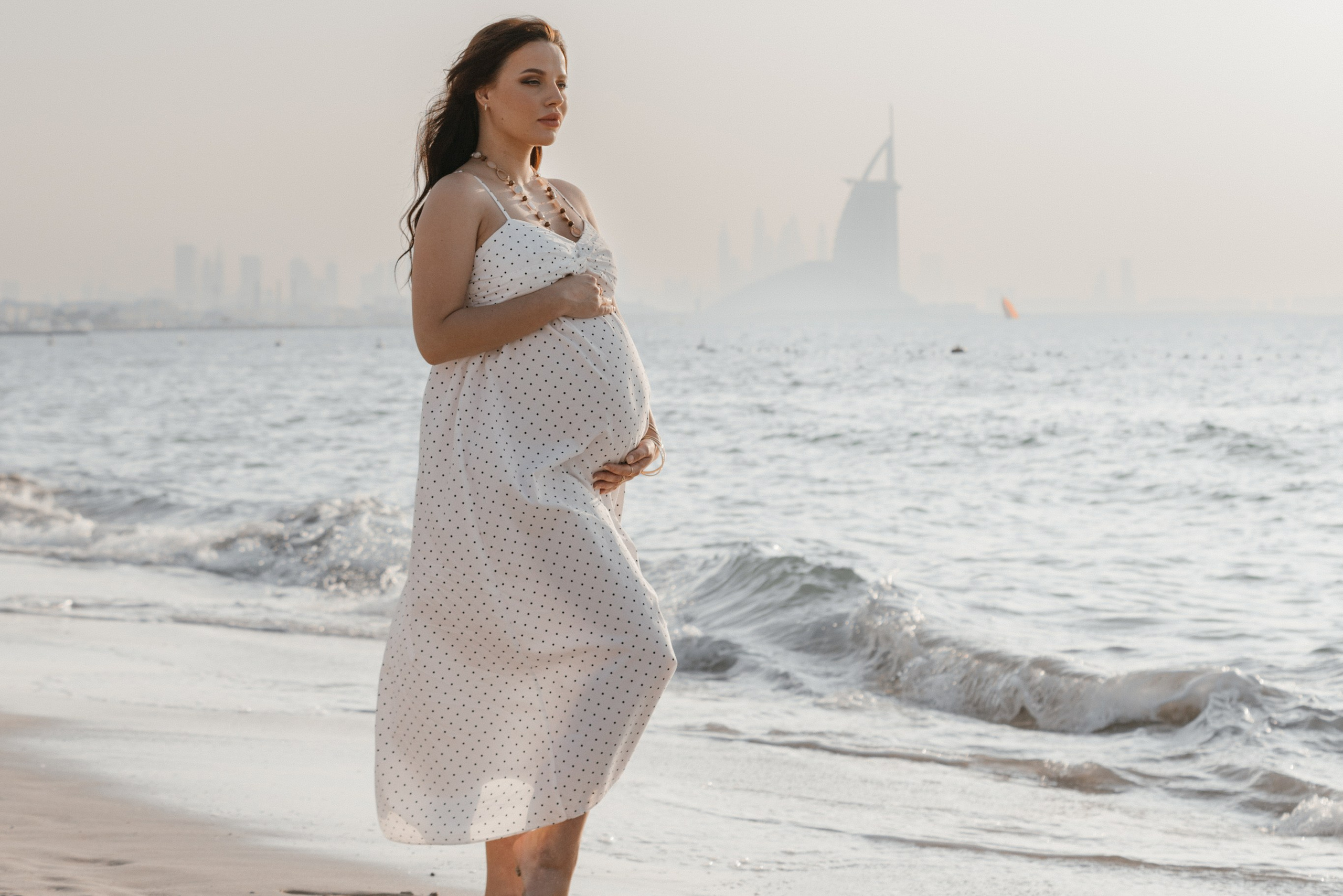 A pregnant woman stands on the beach in Dubai, embracing the tranquility of the sea as the iconic Burj Al Arab is seen in the background