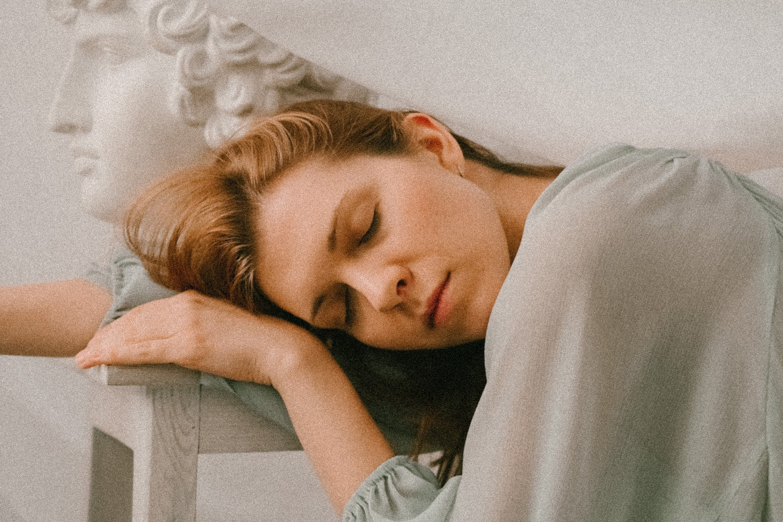 Девушка сидит в струящемся зеленом платье положила голову на руки, рядом гипсовая голова