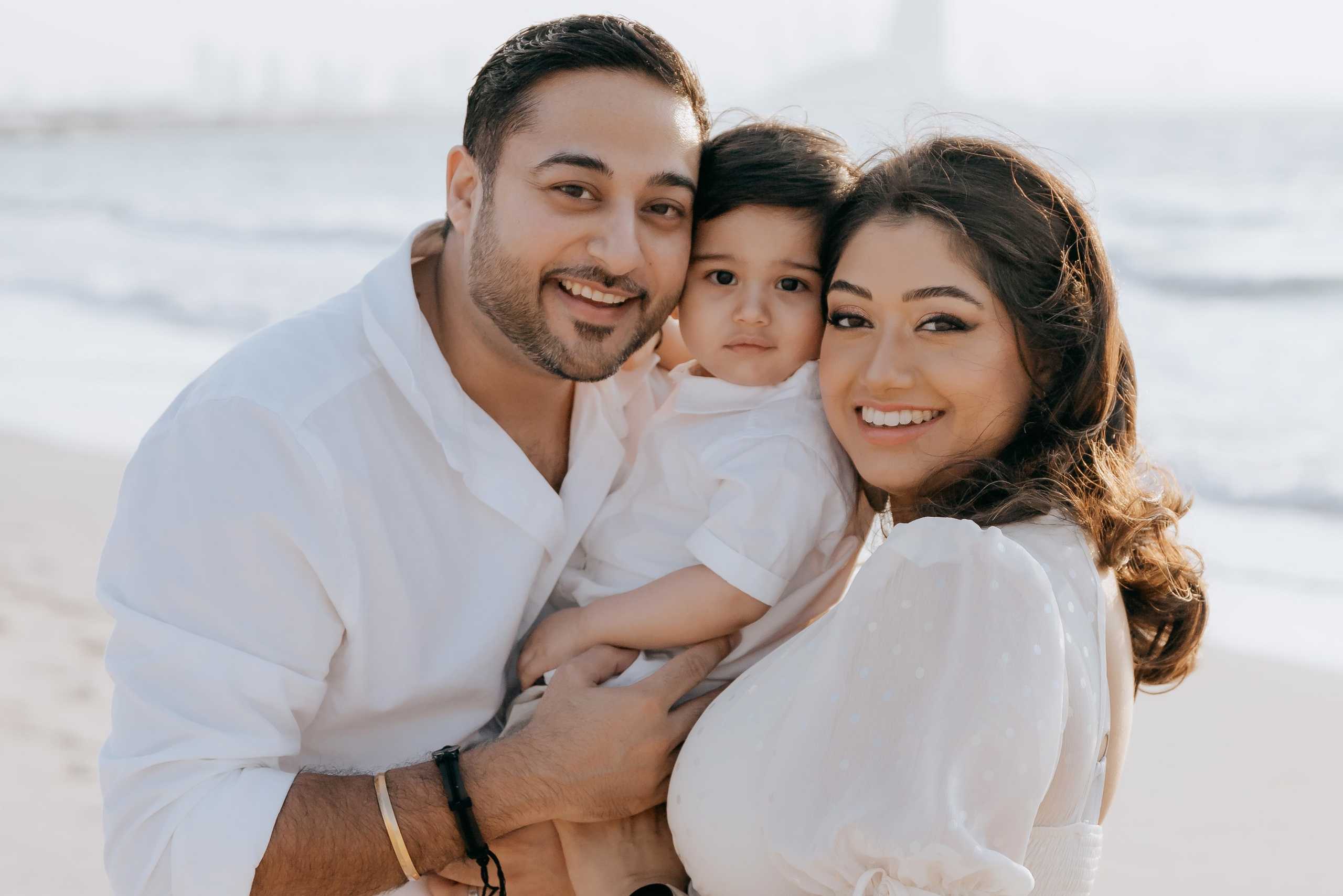 A smiling family enjoys a beach photoshoot in Dubai, holding their young child close with the sea in the background