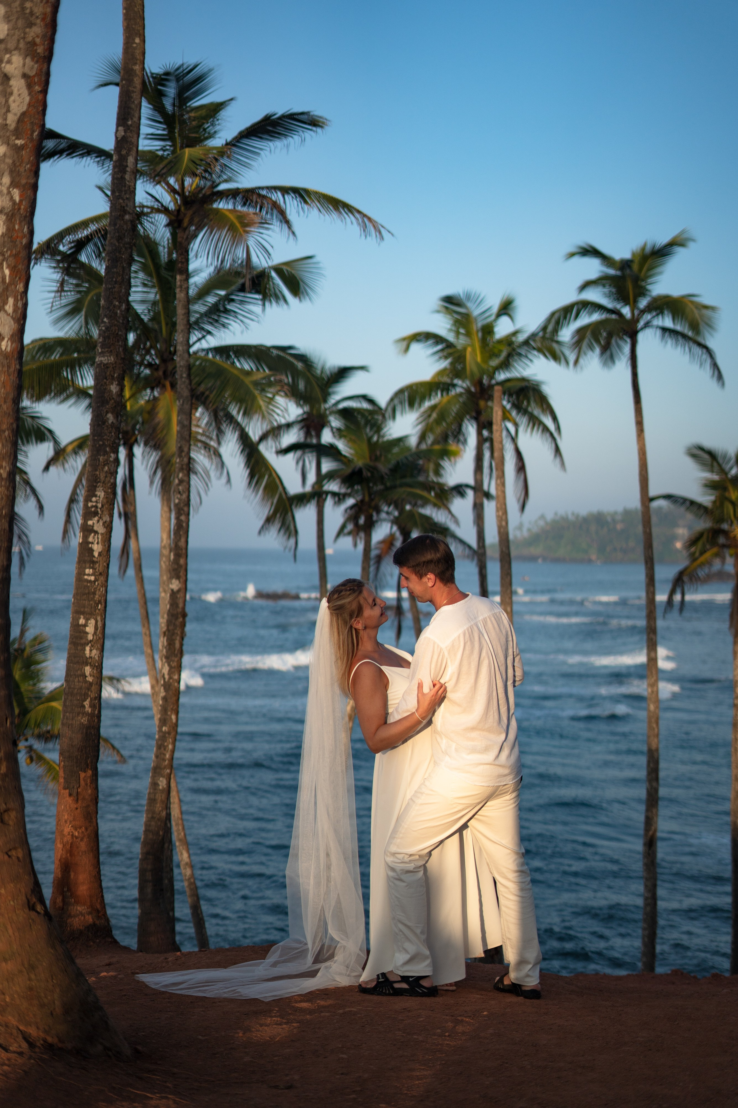 Bride in a wedding dress with a long veil and groom in a white suit posing against the backdrop of Coconut Hill, Sri Lanka