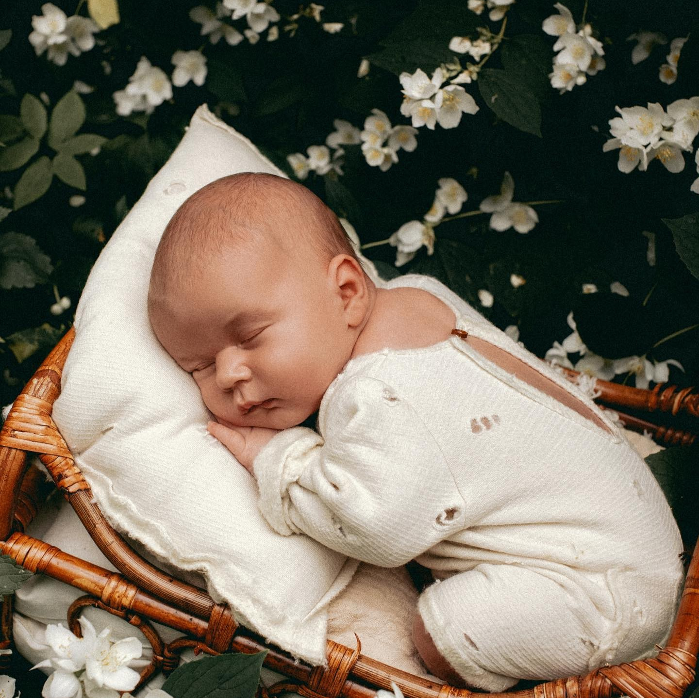 Спящий ребенок в уютной плетеной корзине, окруженный белыми цветами.