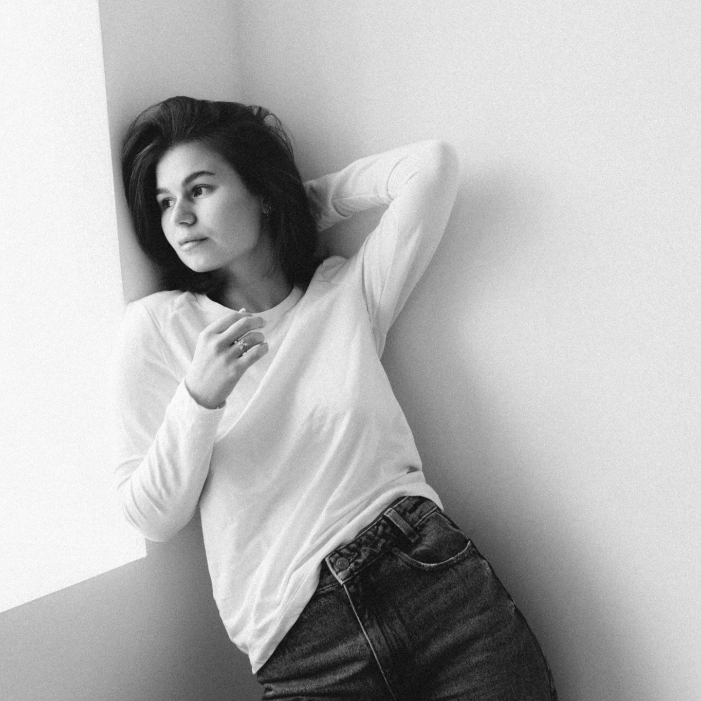 Женщина в белой рубашке с длинными рукавами и джинсах прислонилась к стене, касаясь рукой волос, с задумчивым выражением лица.