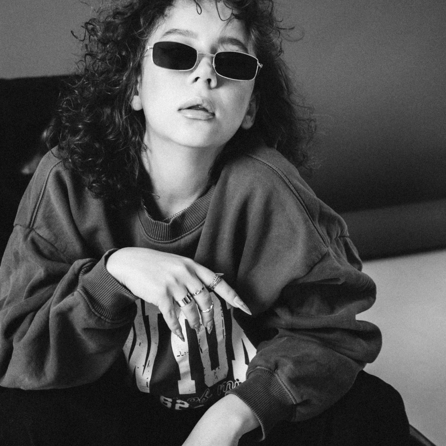 Человек с кудрявыми волосами в солнечных очках и повседневном свитере, сидящий в расслабленной позе.