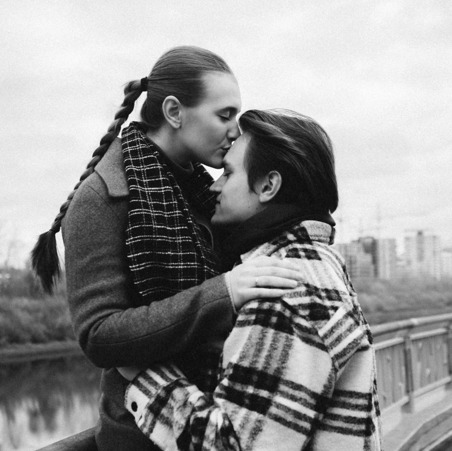 Пара делится сокровенным моментом на мосту, человек слева целует другого в лоб, оба тепло одеты в пальто и шарфы.