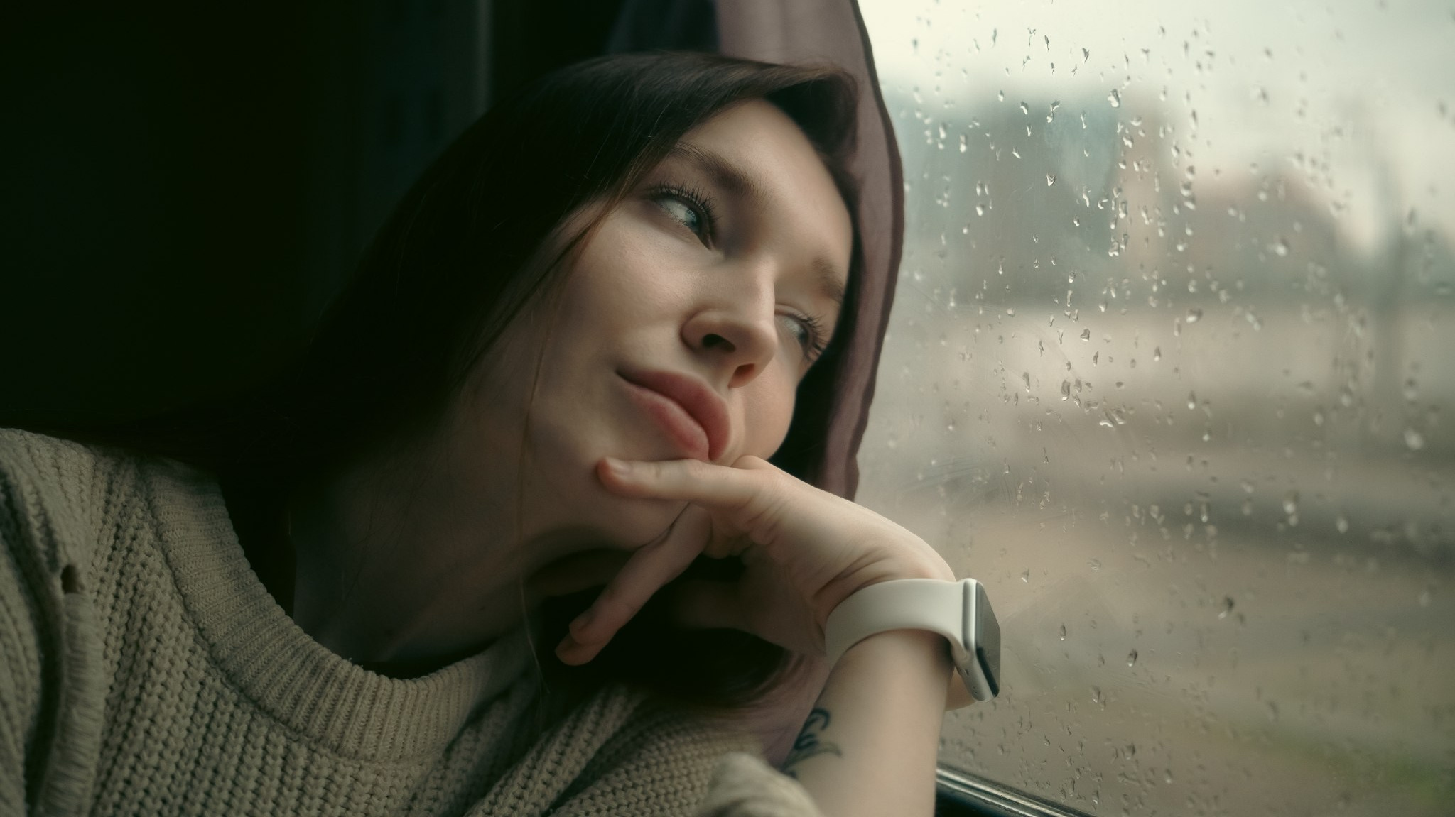 На фотографии изображена молодая девушка, сидящая у окна в транспорте (вероятно, в поезде или автобусе). Она задумчиво смотрит вдаль, слегка приподняв голову и подперев подбородок рукой. Её взгляд кажется мечтательным и немного меланхоличным - будто она погружена в свои мысли или воспоминания. Внешний вид девушки: тёмные волосы свободно спадают на плечи; одета в уютный вязаный свитер светлого оттенка; на запястье - современные смарт-часы с белым ремешком; на предплечье видна татуировка, придающая образу индивидуальность; выражение лица спокойное, с лёгкой грустью или задумчивостью. Обстановка: за окном идёт дождь - стекло покрыто каплями, создающими атмосферу уюта и лёгкой печали; пейзаж за окном размытый, нечёткий, что усиливает ощущение отстранённости от внешнего мира; внутри транспорта полумрак, мягкий свет подчёркивает черты лица девушки и текстуру её одежды; рядом с девушкой виден фрагмент занавески или шторки, что добавляет интимности сцене. Общее настроение снимка: Фотография передаёт атмосферу уединения, размышлений и лёгкой грусти. Сочетание дождливой погоды за окном, задумчивого взгляда девушки и приглушённого освещения создаёт ощущение ностальгии и внутренней глубины. Снимок может ассоциироваться с моментом личного переосмысления, ожидания или мечтаний о чём-то далёком и важном.