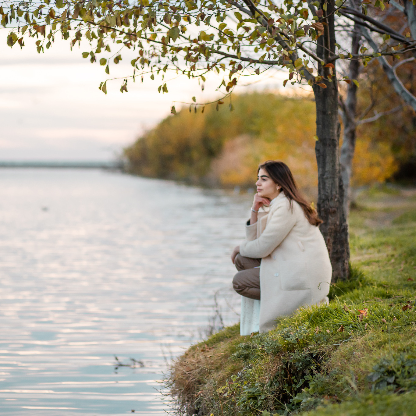 Девушка присела у берега реки на фоне осеннего леса и смотрит в даль