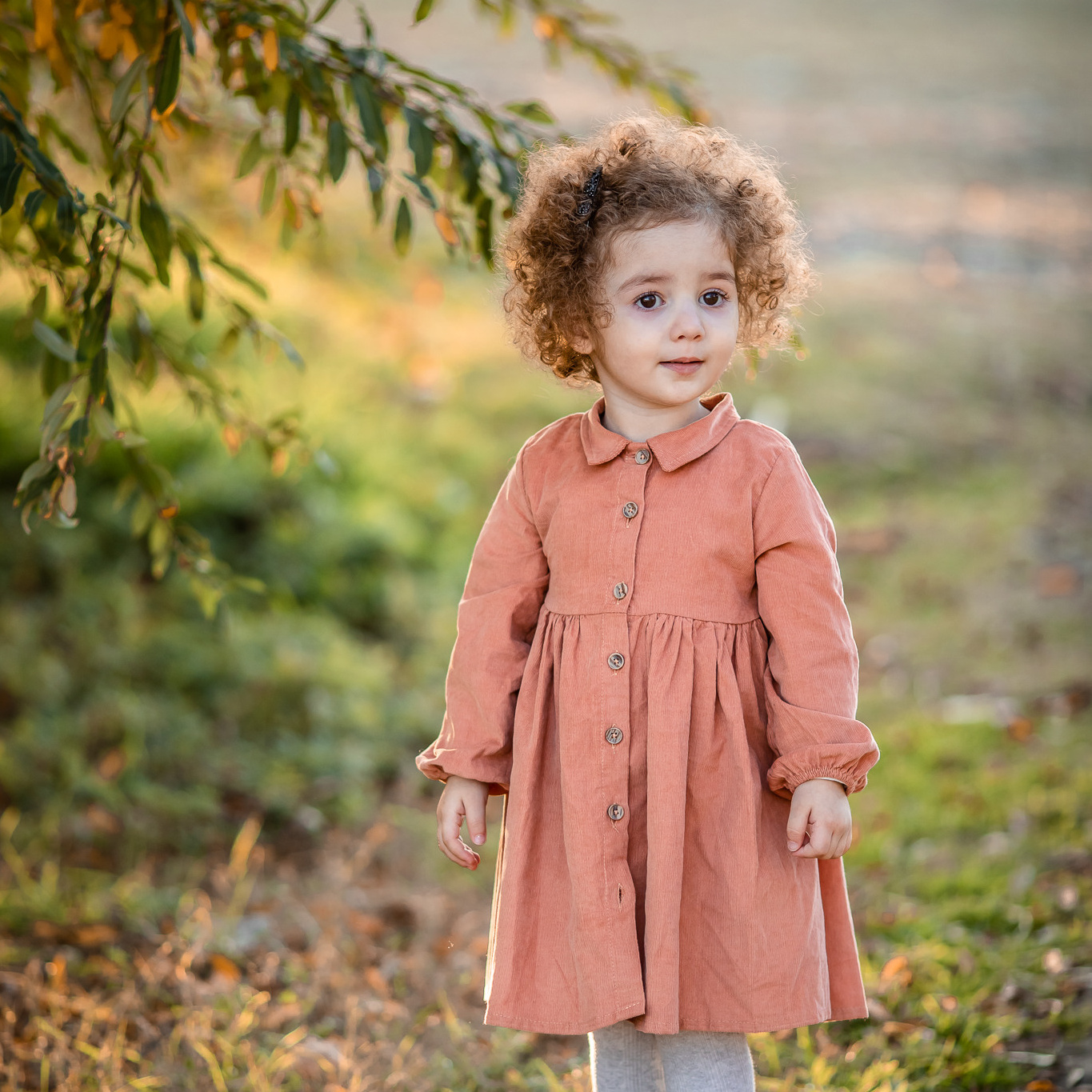 Маленькая кудрявая девочка в платье персикового цвета стоит возле зеленных деревьев 