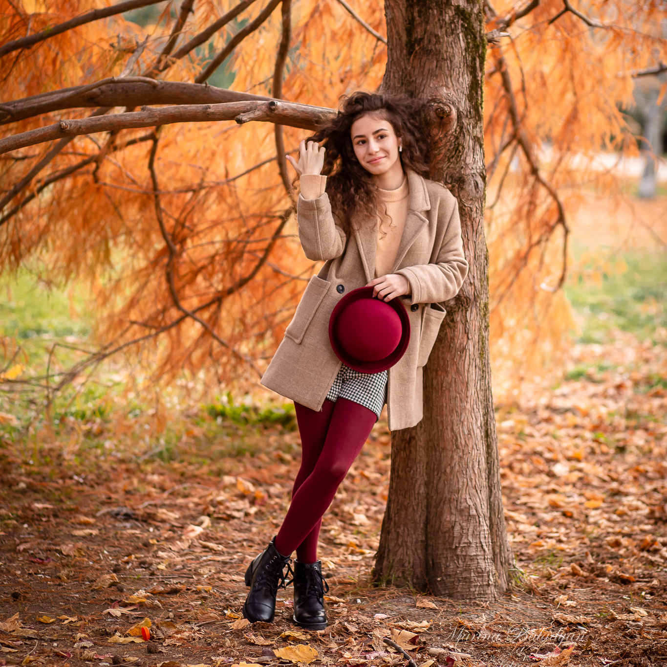 Красивая девушка в бордовых колготках стоит у осеннего дерева среди листвы.