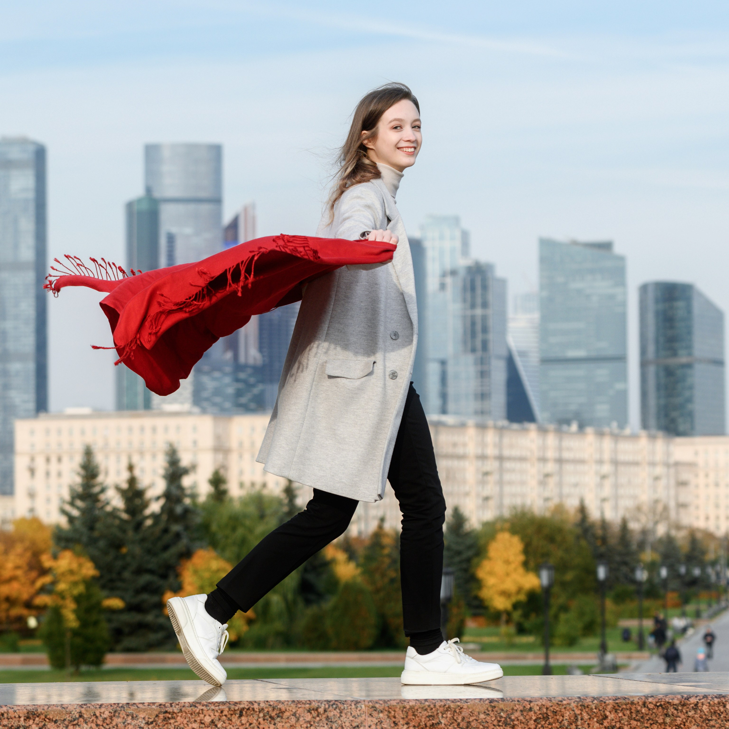 Девушка в движении подбрасывает красный шарф на фоне городских высоток и осенних деревьев.