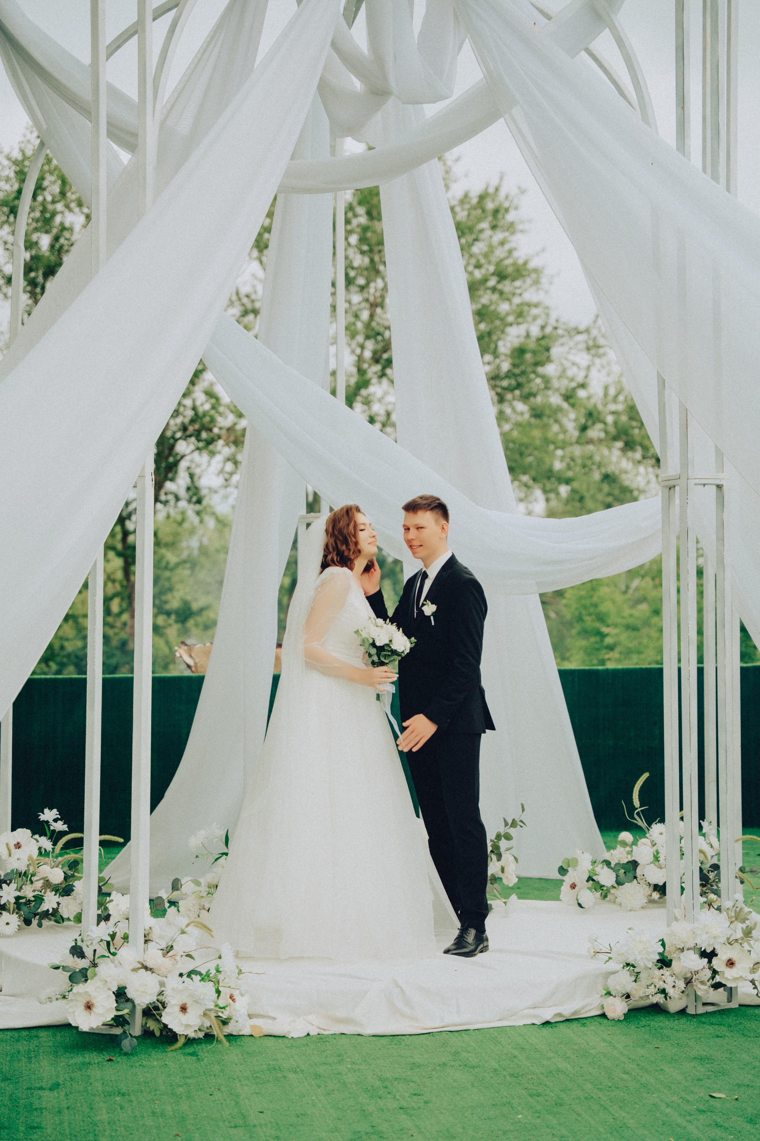 Момент перед ЗАГСом (Волнение, ожидание)Невеста у зеркала в комнате ожидания: Невеста с волнением поправляет фату в зеркале комнаты ожидания ЗАГСа, за секунду до начала церемонии.Жених ждет у дверей: Жених nervously проверяет время у дверей свадебного зала, в ожидании своей невесты.Родители невесты: Отец невесты с гордостью и слезами на глазах смотрит на дочь перед выходом.2. Церемония (Кульминация)Вход невесты: Двери зала открываются, и невеста в ослепительном платье делает первый шаг к жениху под взгляды гостей.Обмен клятвами: Крупный план: жених, держа за руки невесту, с серьезным и loving взглядом произносит誓言 (клятву).Слезы счастья: Слеза радости скатывается по щеке невесты в момент произнесения торжественных слов.Взгляд со стороны: Молодожены стоят у арки, слушая речь сотрудника ЗАГСа, их руки крепко сплетены.Реакция гостей: Гости ceremoniously наблюдают за церемонией, на их лицах — улыбки и умиление.3. Обмен кольцамиКрупный план рук: Дрожащие руки жениха надевают обручальное кольцо на палец невесты. Крупный план, свадебная фотография.Эмоции в момент: Невеста заливается счастливым смехом, пытаясь надеть кольцо жениху.4. Первый поцелуй как супруговКульминационный момент: Первый поцелуй молодоженов как муж и жена под аплодисменты гостей в свадебном зале ЗАГСа.Крупный план: Невеста и жених целуются на фоне яркого света окон и размытых силуэтов гостей.5. После церемонии (Облегчение, радость)Выход из зала: Новые муж и жена с сияющими улыбками выходят из зала ЗАГСа под рис и аплодисменты.Объятия с родителями: Невеста нежно обнимает свою маму сразу после окончания церемонии, обе счастливы.Поздравления гостей: Друзья бросаются обнимать молодоженов в фойе ЗАГСа, все смеются и поздравляют.6. Работа с документамиПодписание: Молодожены вместе ставят подписи в официальном свадебном свидетельстве, этот момент навсегда в истории их семьи.Получение паспортов: Жених с улыбкой получает из рук сотрудницы ЗАГСа паспорт со штампом о браке.