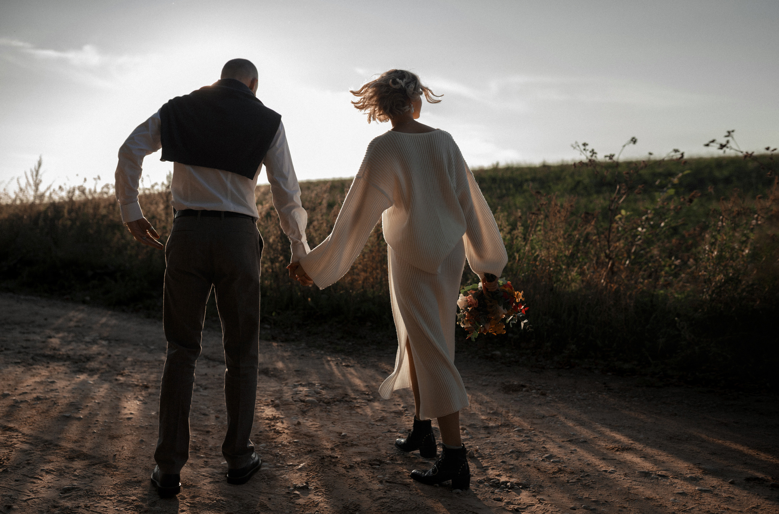 Взявшись за руки пара новобрачных идет по пыльной дороге напротив к солнцу.