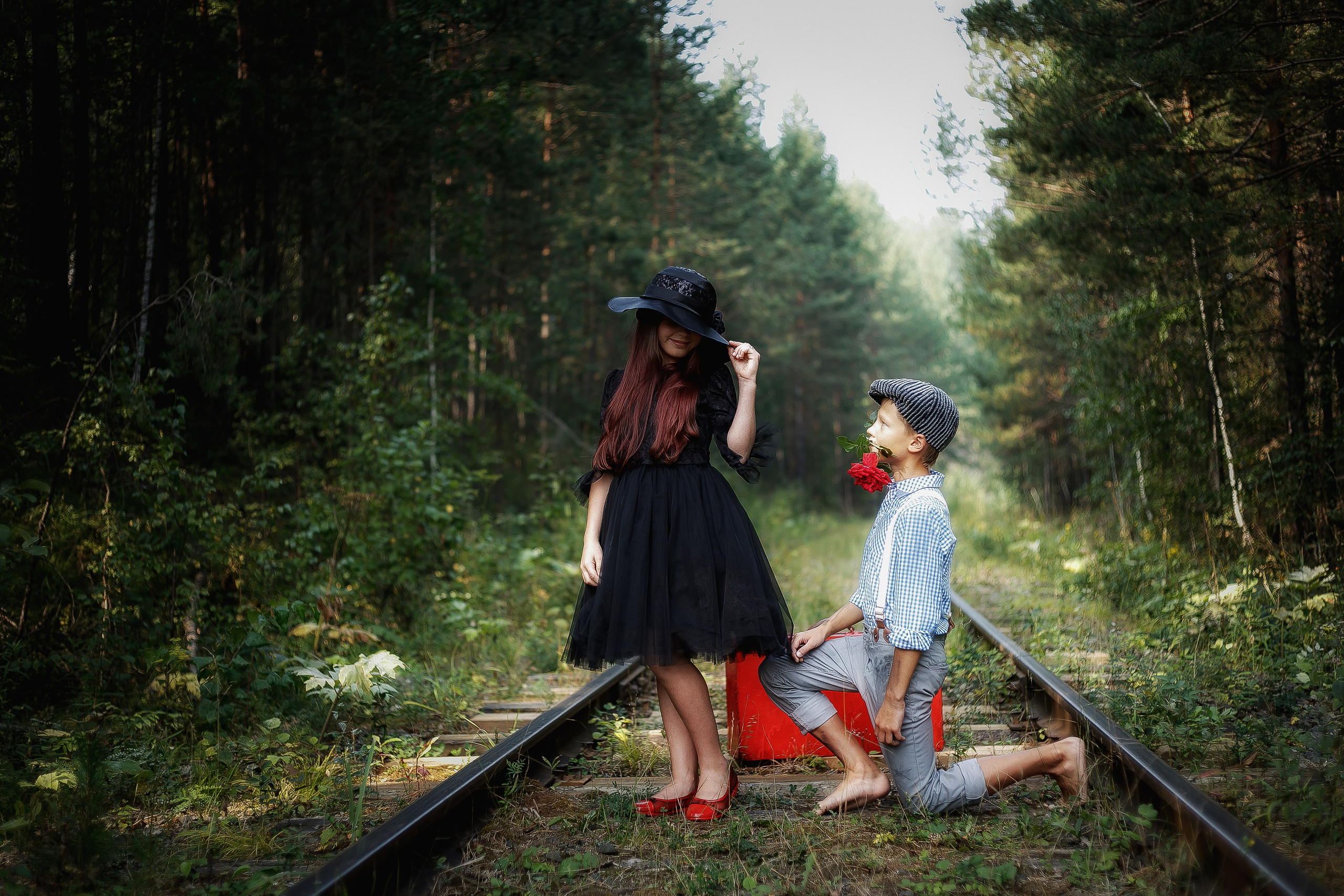 Мальчик на коленях с красной розой во рту делает предложение девочке в черном платье и шляпе, они стоят на заброшенных железнодорожных путях в лесу.