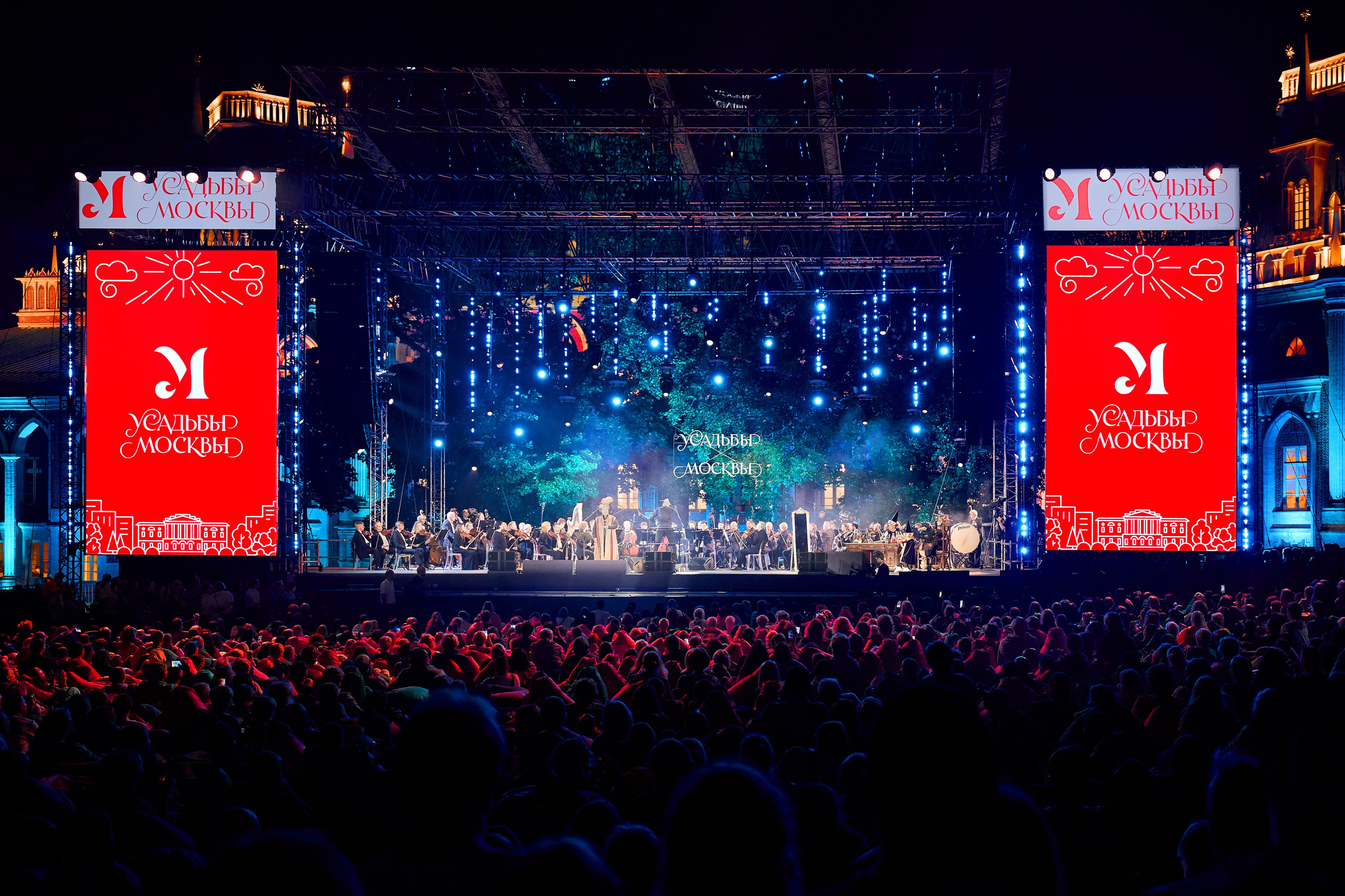 Фотосъёмка большого концерта в парке-усадьбе Царицыно для проекта 'Московские усадьбы'. На сцене оркестр под яркими огнями, а в центре — большое изображение логотипа проекта 'Московские усадьбы'. Зрители внизу, освещённые красными огнями, наслаждаются выступлением. Вокруг сцены — красивое ночное освещение и большие экраны с визуальными эффектами