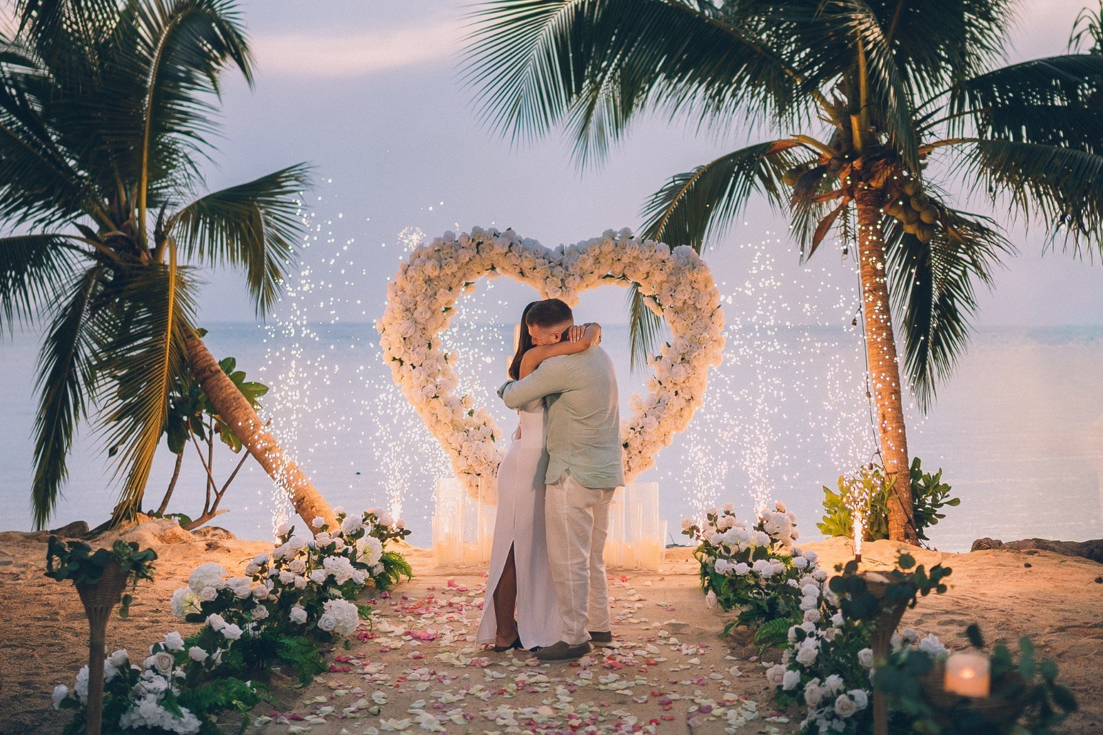 Romantic beach proposal in Koh Samui with a couple under palm trees, white flowers and sparkling fireworks at night.