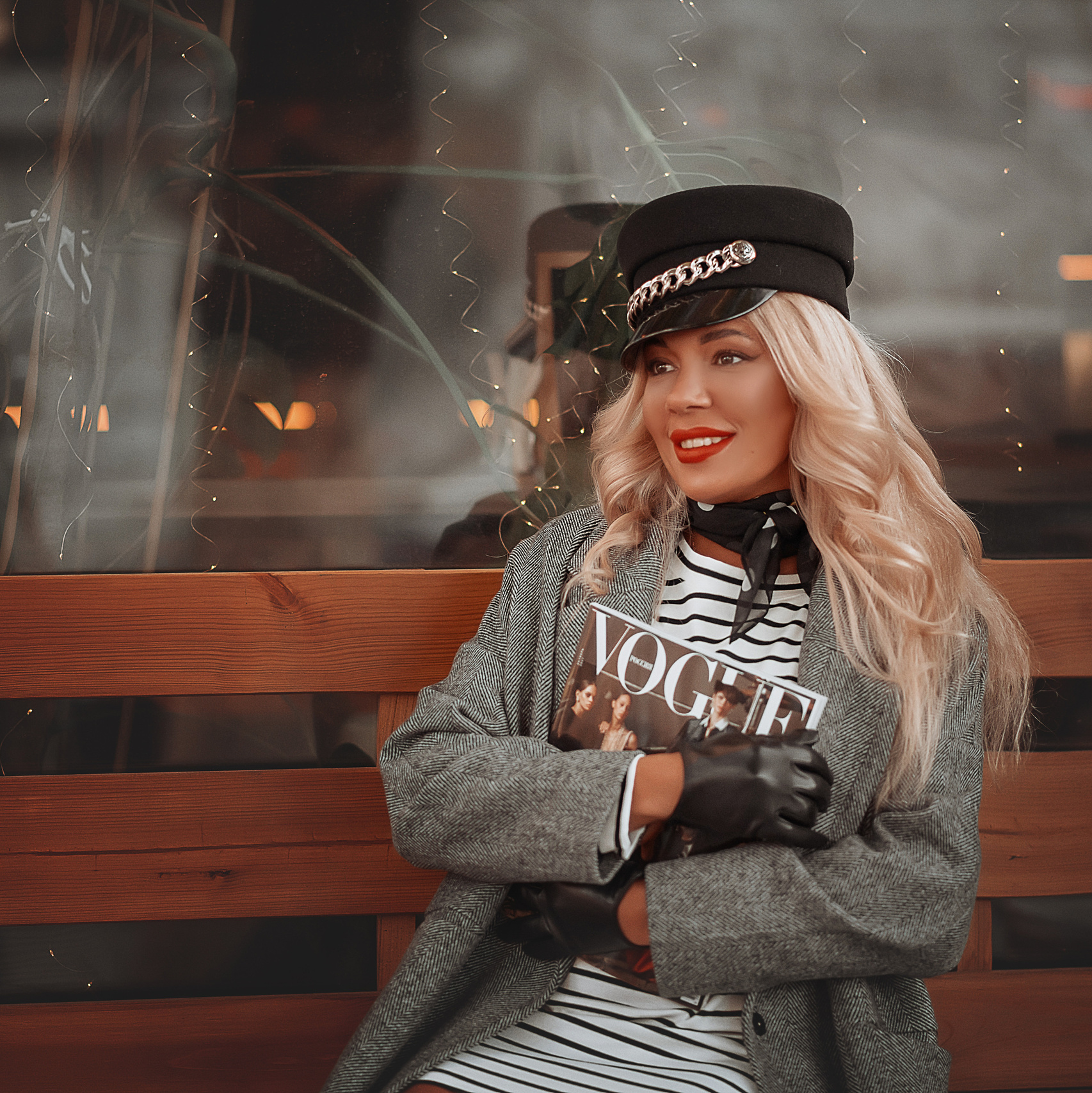 Девушка в кепке и пальто сидит на скамейке с журналом в руках.