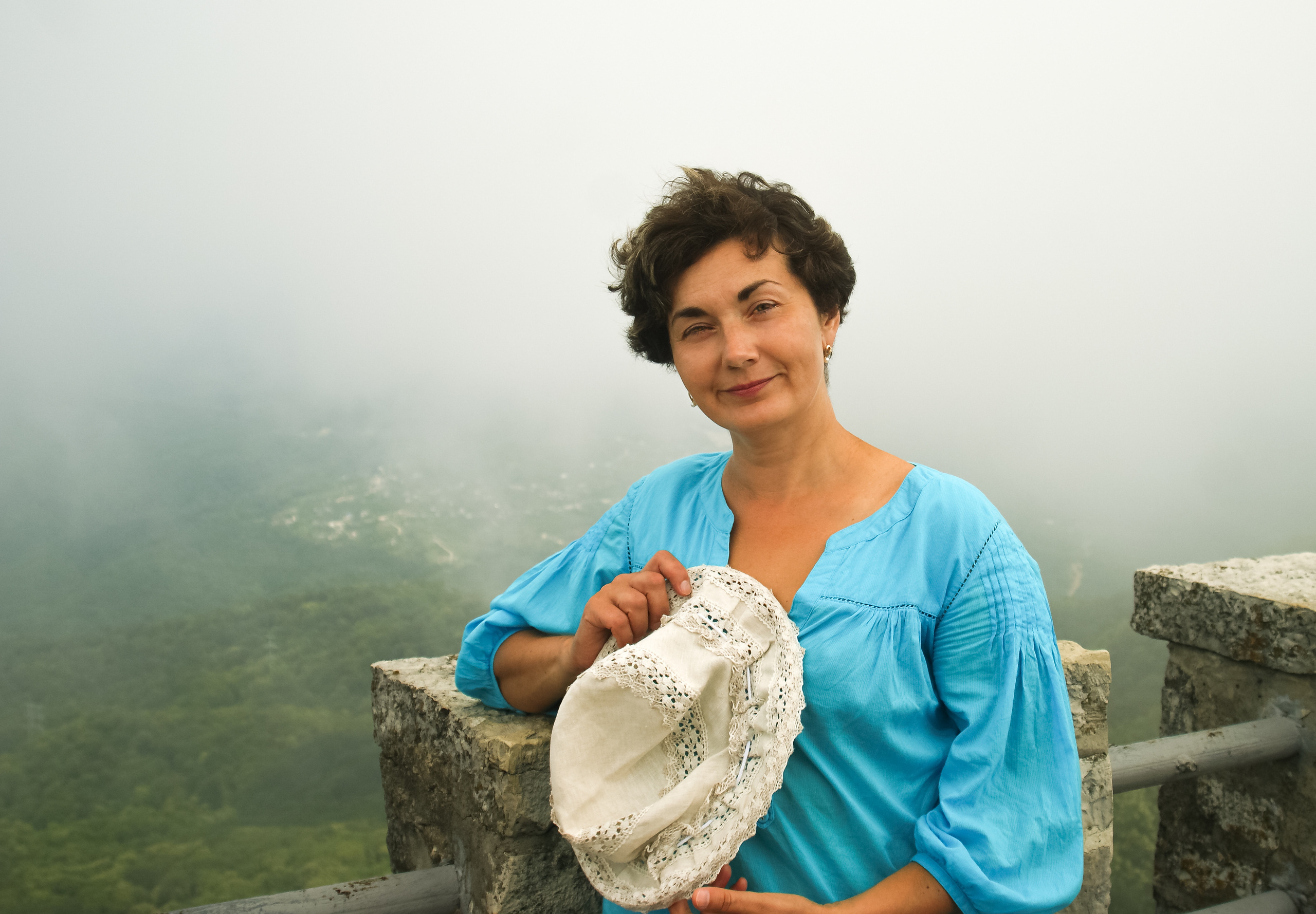 Женщина в голубой блузке со шляпой в руке на фоне тумана.