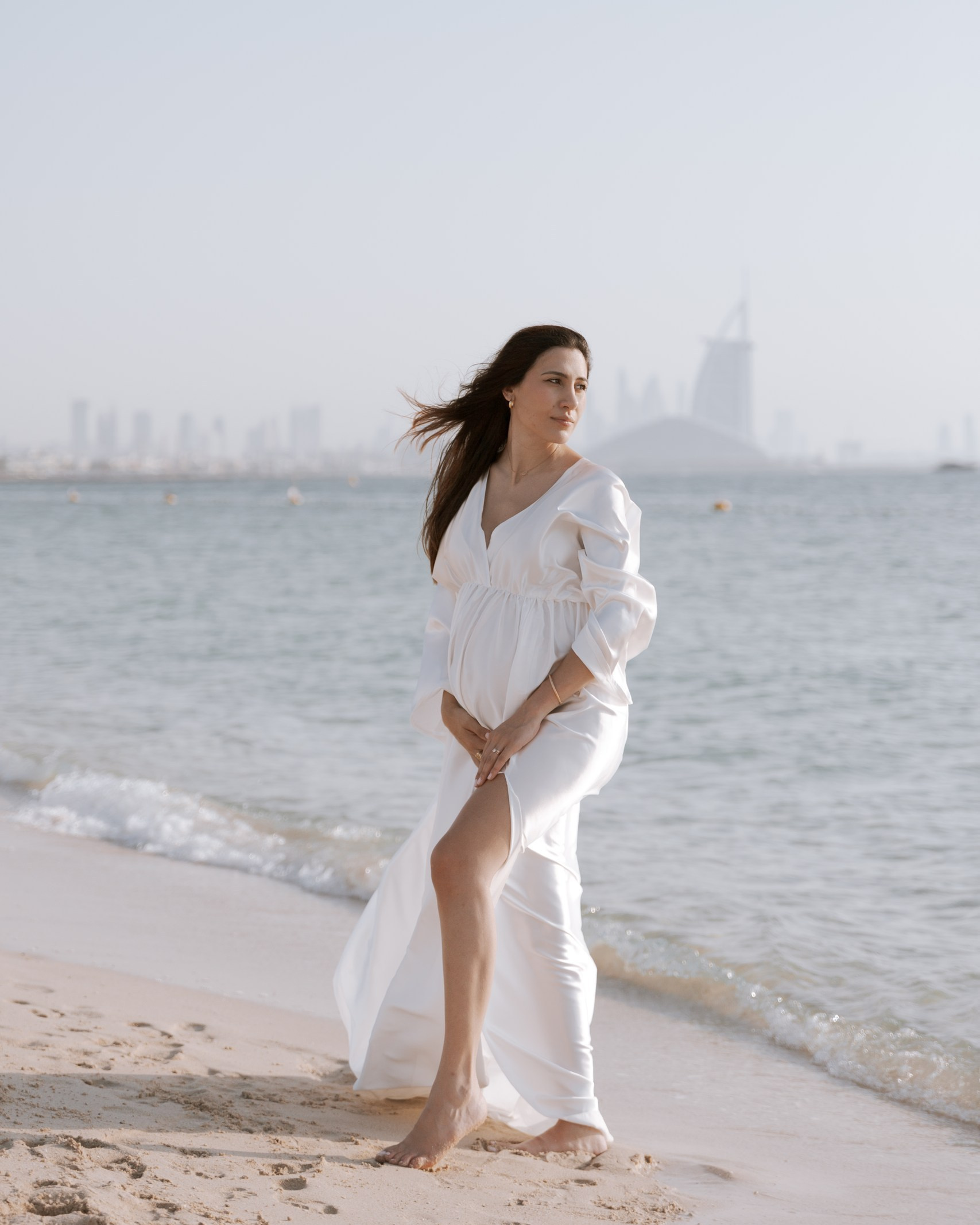 Pregnant woman in a white dress standing on a beach, capturing the best maternity photo in Emirates with the ocean and city skyline in the background