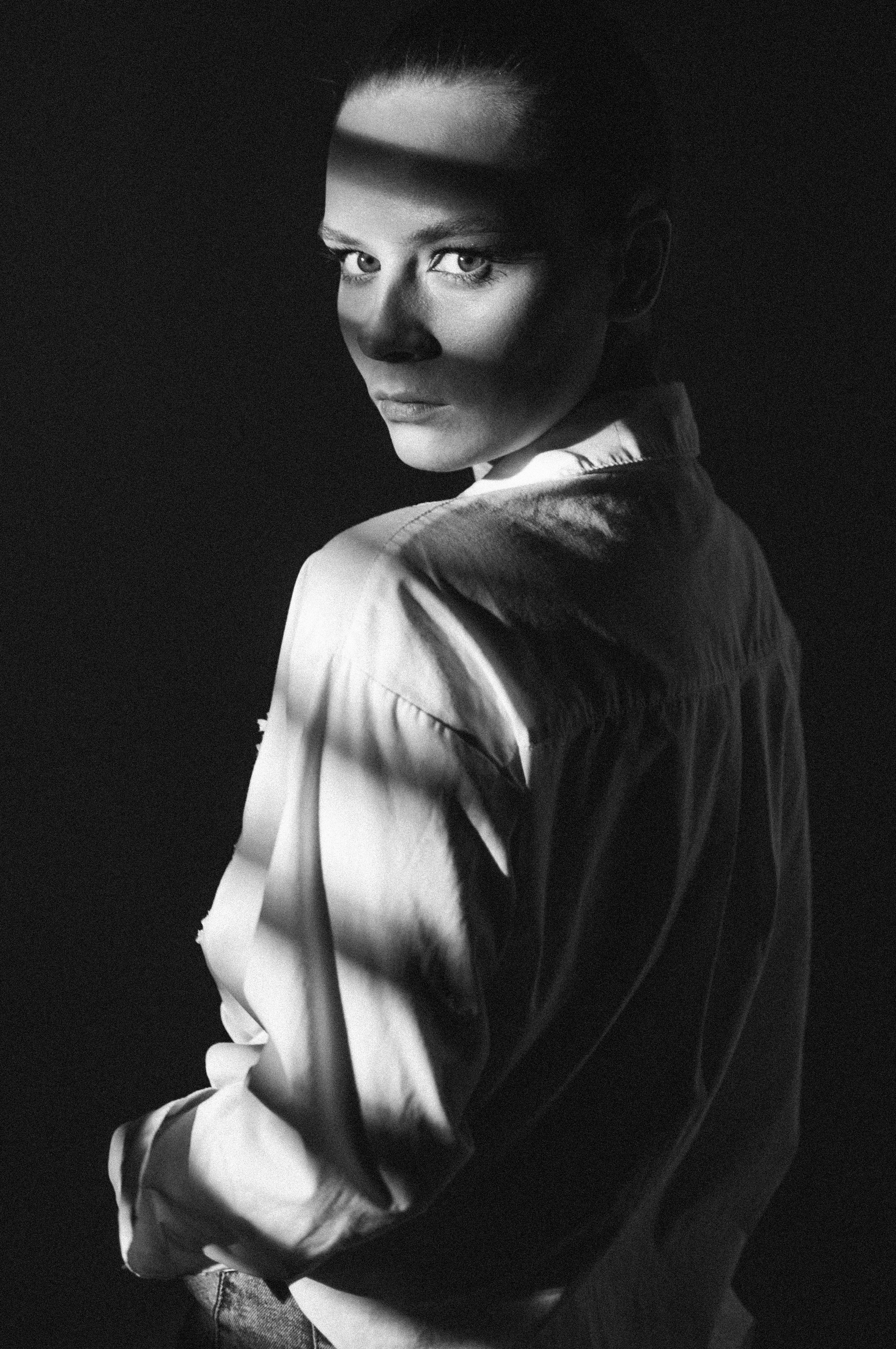 Черно-белый портрет девушки на темном фоне в нуар-стиле. Свет пробивается на ее лицо через жалюзи.