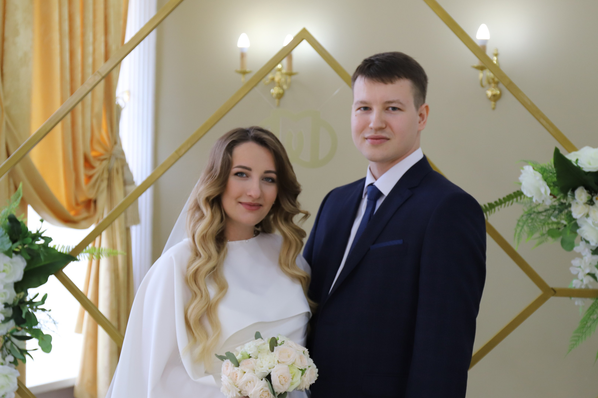 Молодожены в зале торжественных церемоний. Невеста в белом платье держит букет белых роз, рядом стоит жених в темно-синем костюме. Классический свадебный портрет молодых. Элегантный интерьер с золотистыми элементами и белыми цветами.