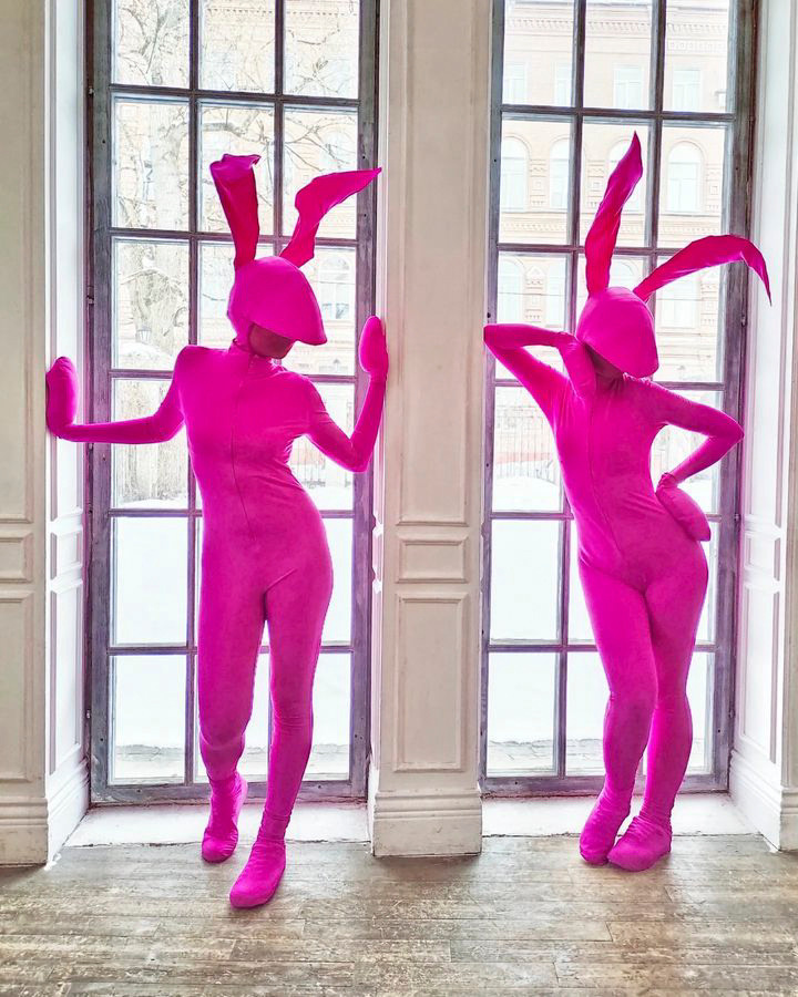 Артисты девушки в обтягивающих костюмах зайчиков цвета фуксии стоят у окна.