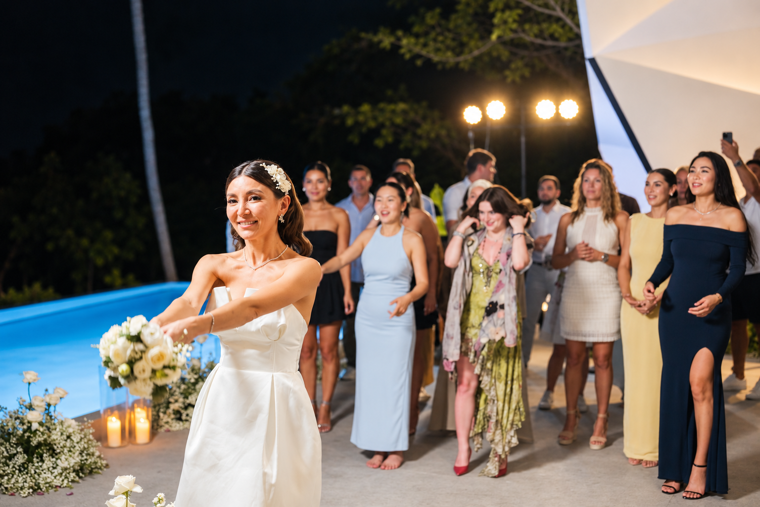 Bride tossing her wedding bouquet to a group of excited female guests during an outdoor evening wedding reception by the pool in Koh Samui, Thailand.