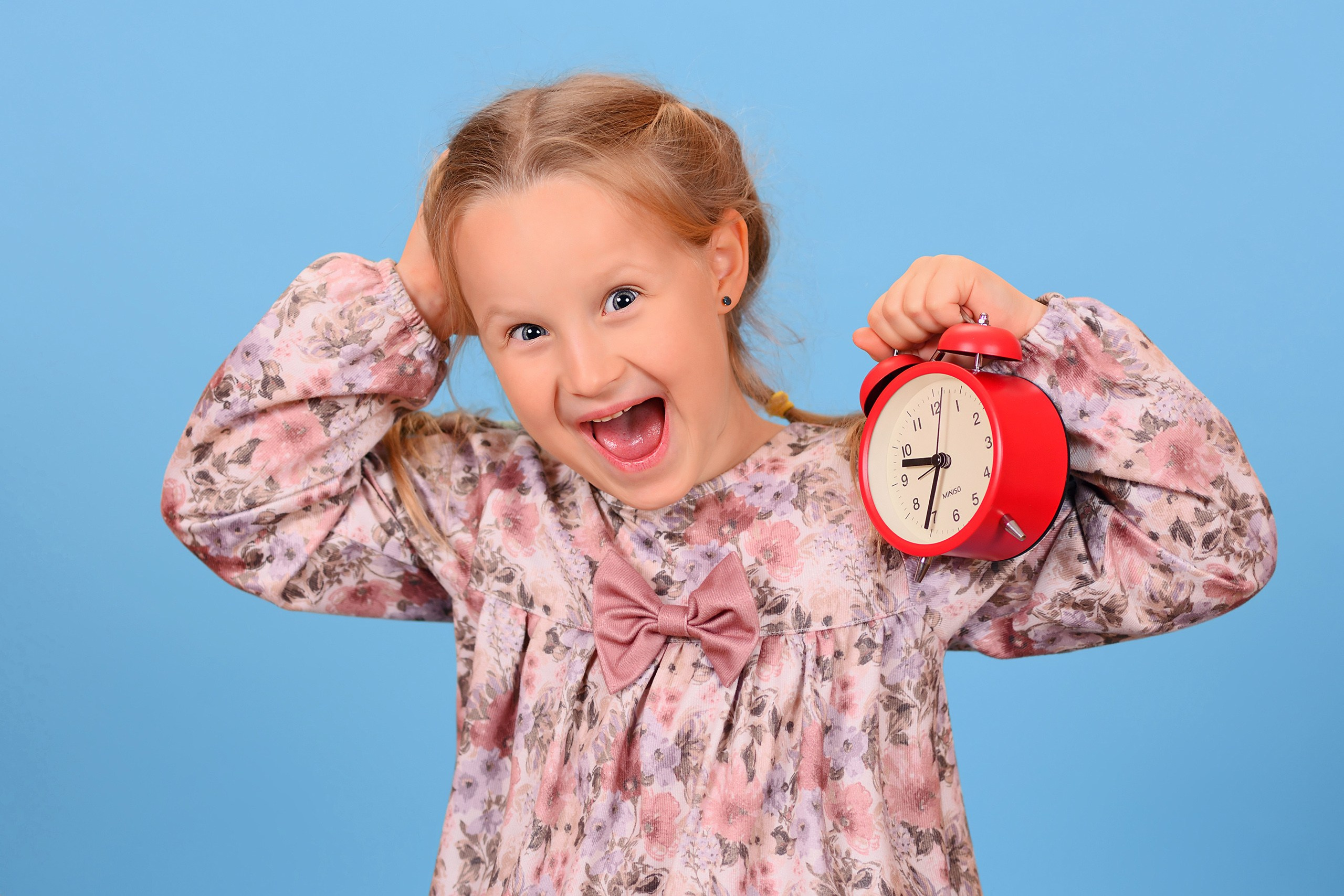 Эмоциональный портрет девочки в детском саду с будильником в руках, изображающей, что она проспала.