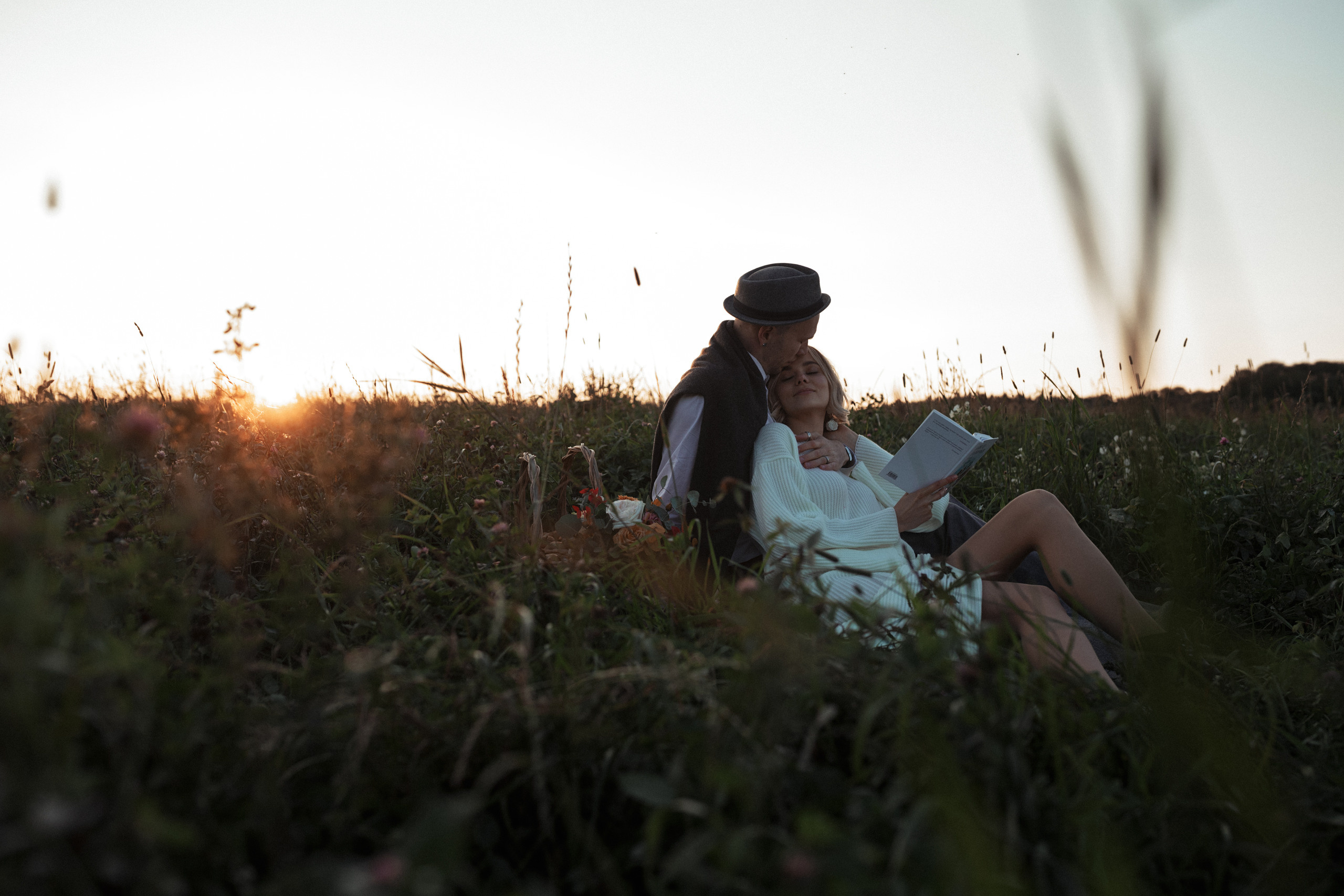 Пара молодоженов лежат в траве в поле. Невеста прислонилась спиной к жениху, он читает ей книгу.