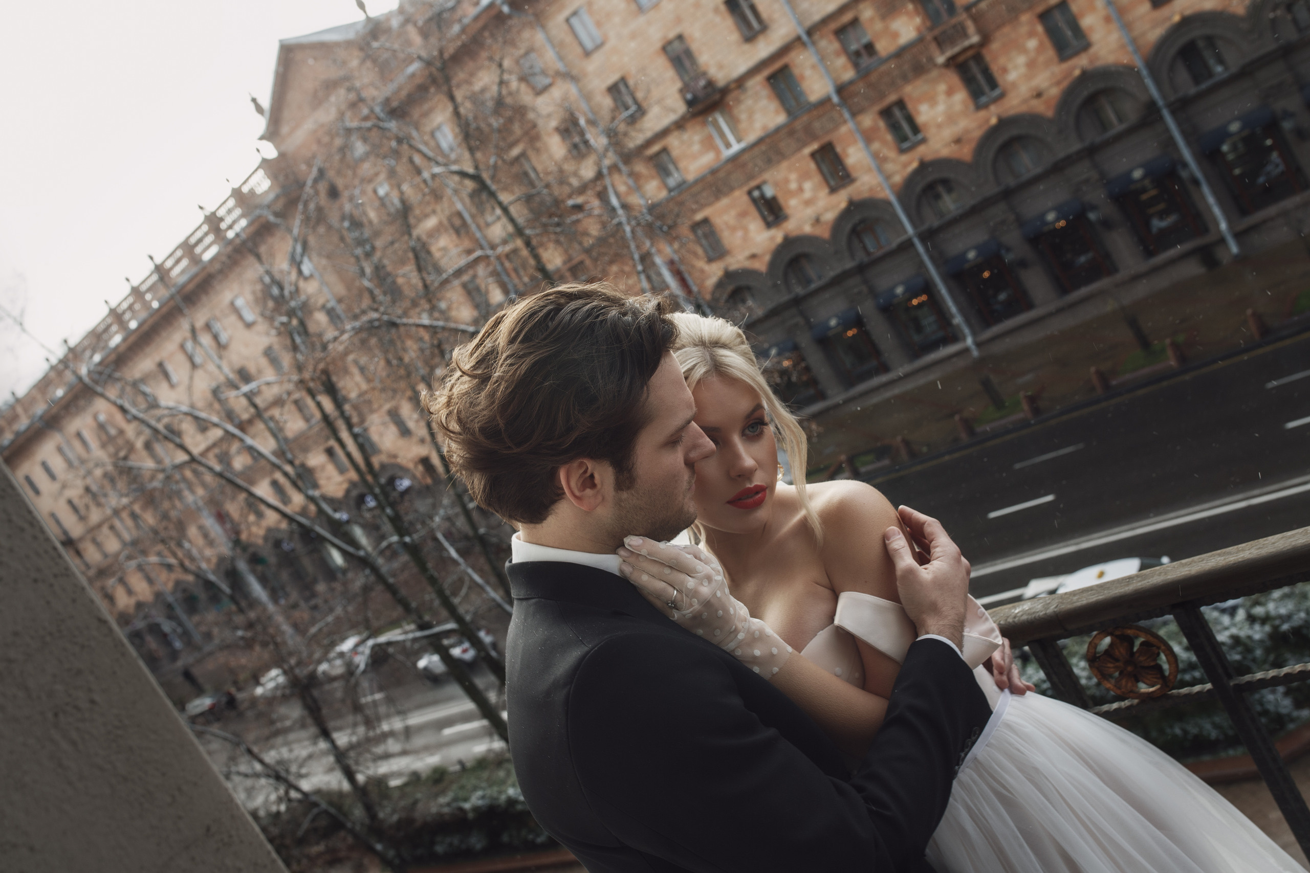 Жених обнимает свою невесту. Берет ее руки в белых перчатках в свои и целует.